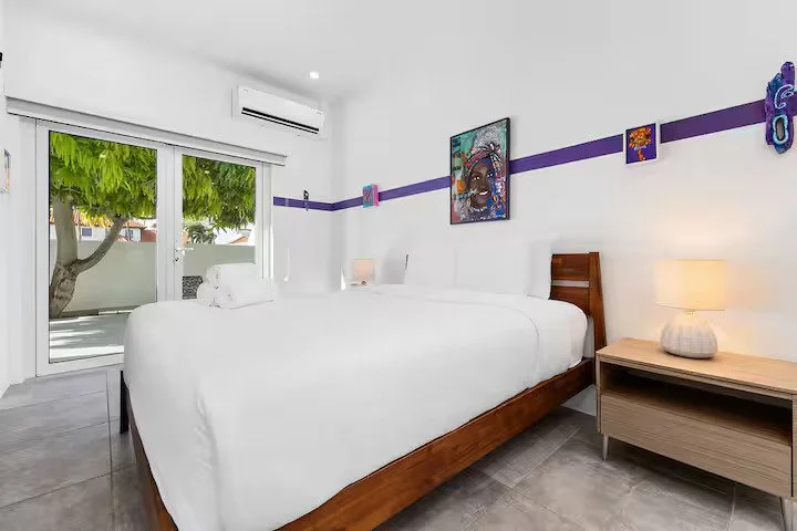 Bedroom with a large bed, wooden headboard, white bedding, bedside lamps, and wall decorations, with sliding glass door leading to a balcony with green trees outside.