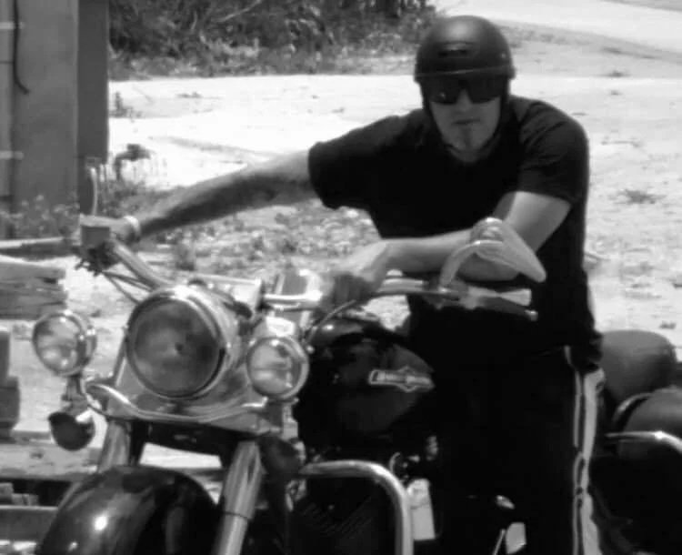 A person wearing a helmet and sunglasses sitting on a motorcycle outdoors in a black-and-white photo.