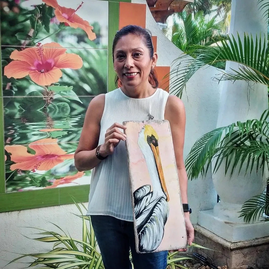 A woman standing outdoors, smiling, holding a painting of a pelican in front of a tropical-themed background with large green plants and flower pictures.