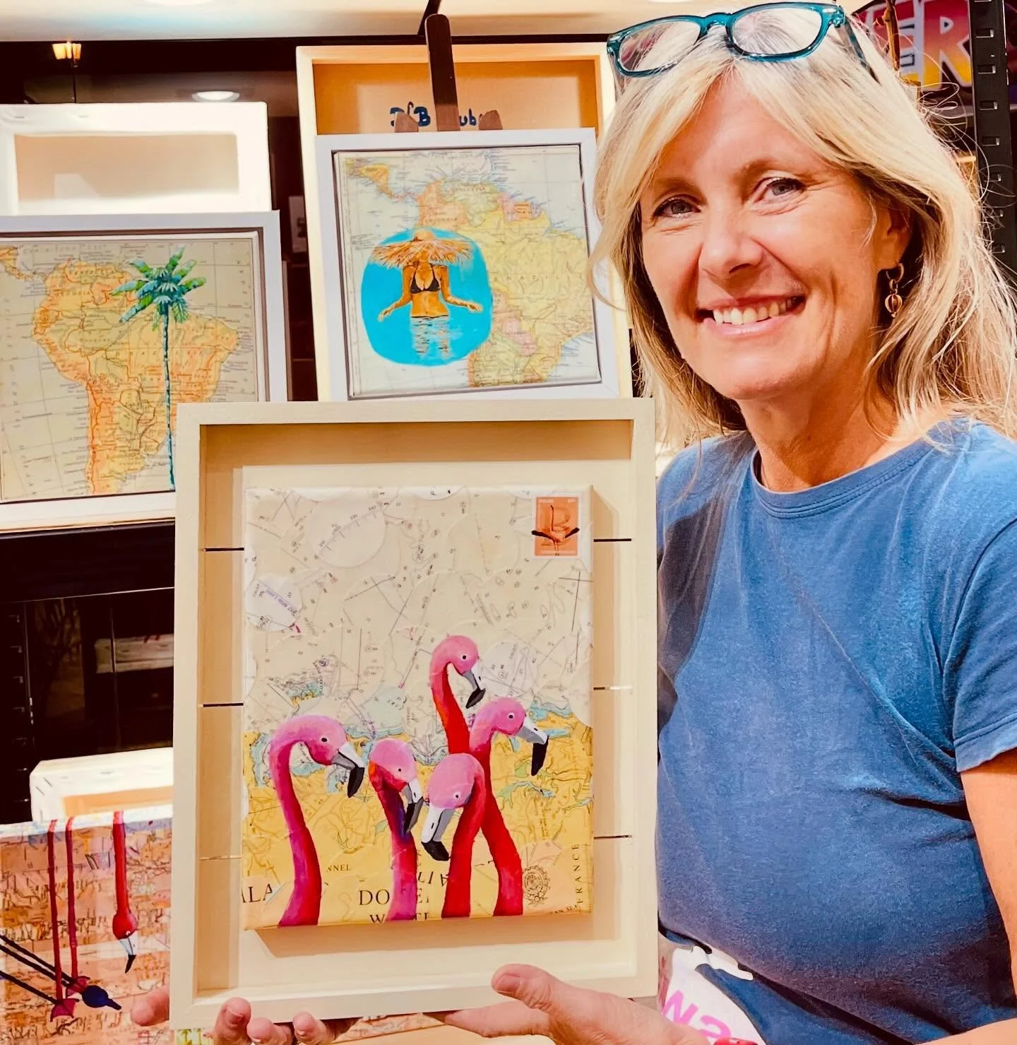 A woman smiling and holding an art piece of five pink flamingos made from painted wooden figures, set against a background of maps with tropical themes. The woman has blonde hair, wears glasses on her head, and a blue t-shirt.