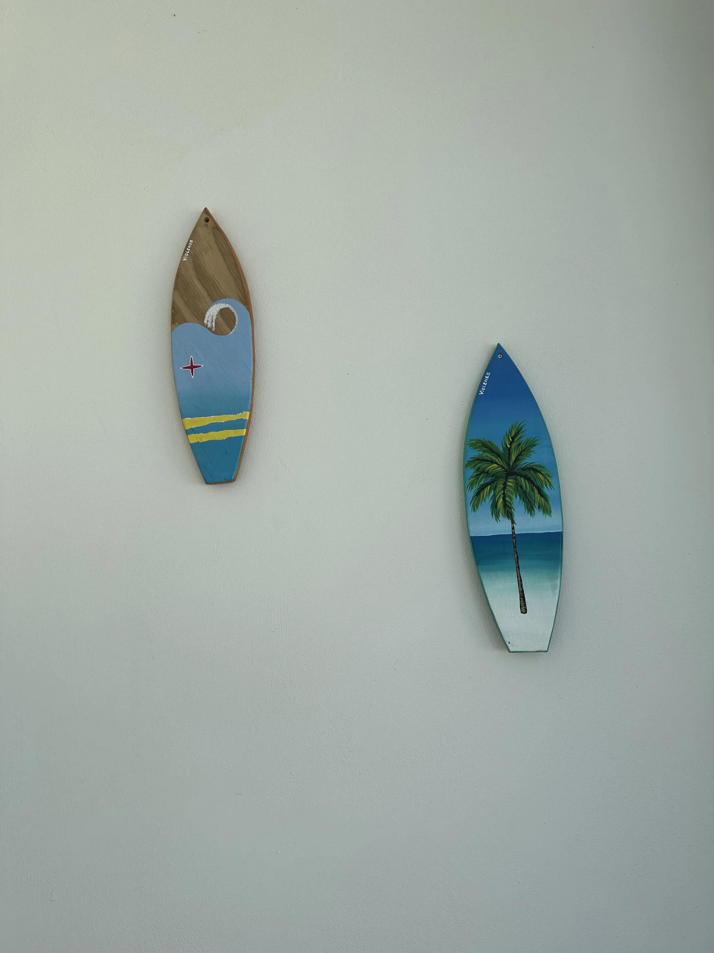 Two painted surfboards hanging on a plain white wall. The left surfboard is painted with a blue wave and yellow stripes, and the right surfboard features a painted image of a palm tree on a beach scene.