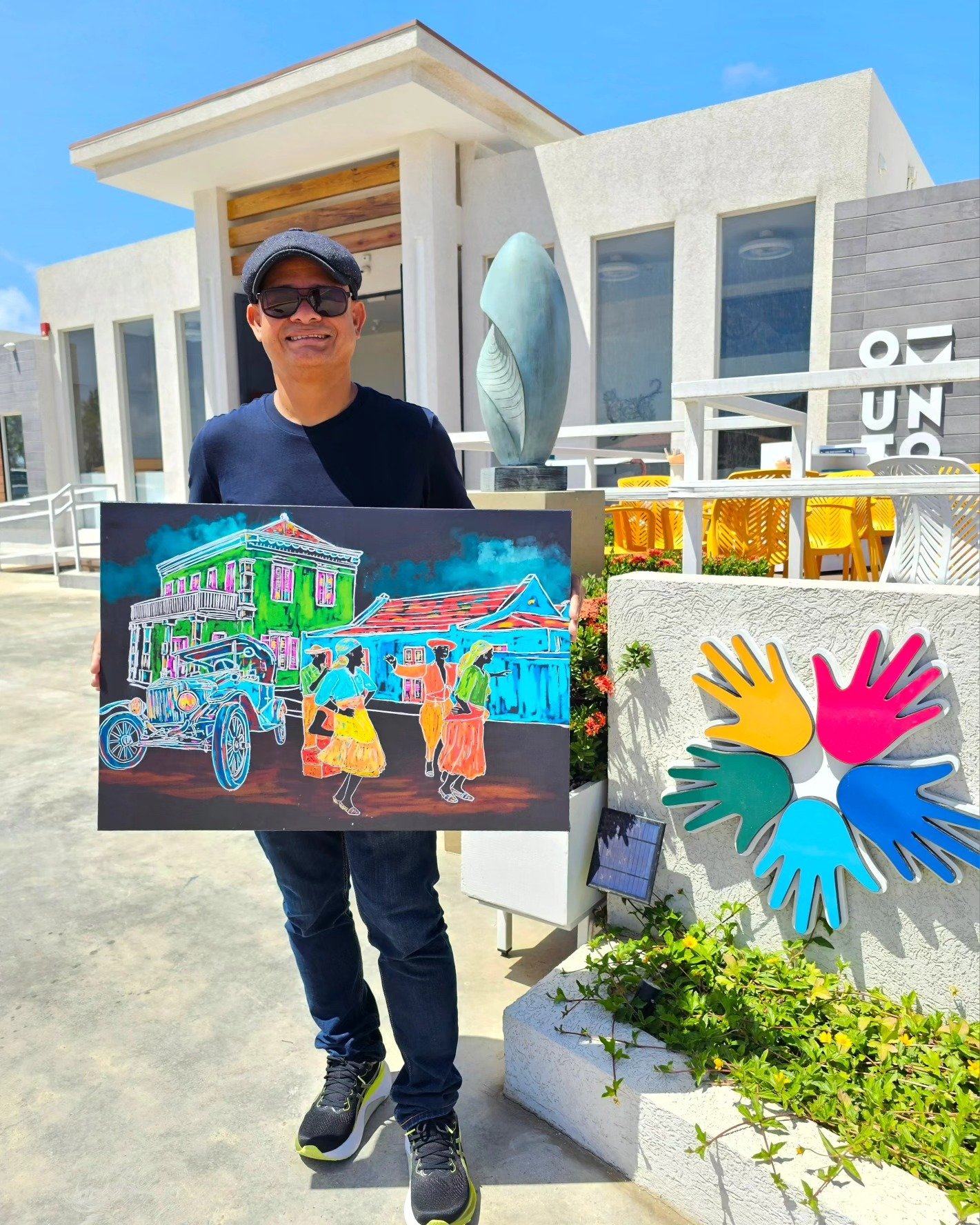 A man wearing sunglasses and a cap holding a colorful painting in front of a modern building with a sculpture, yellow chairs, and a vibrant logo of handprints. The scene is outdoors on a sunny day.