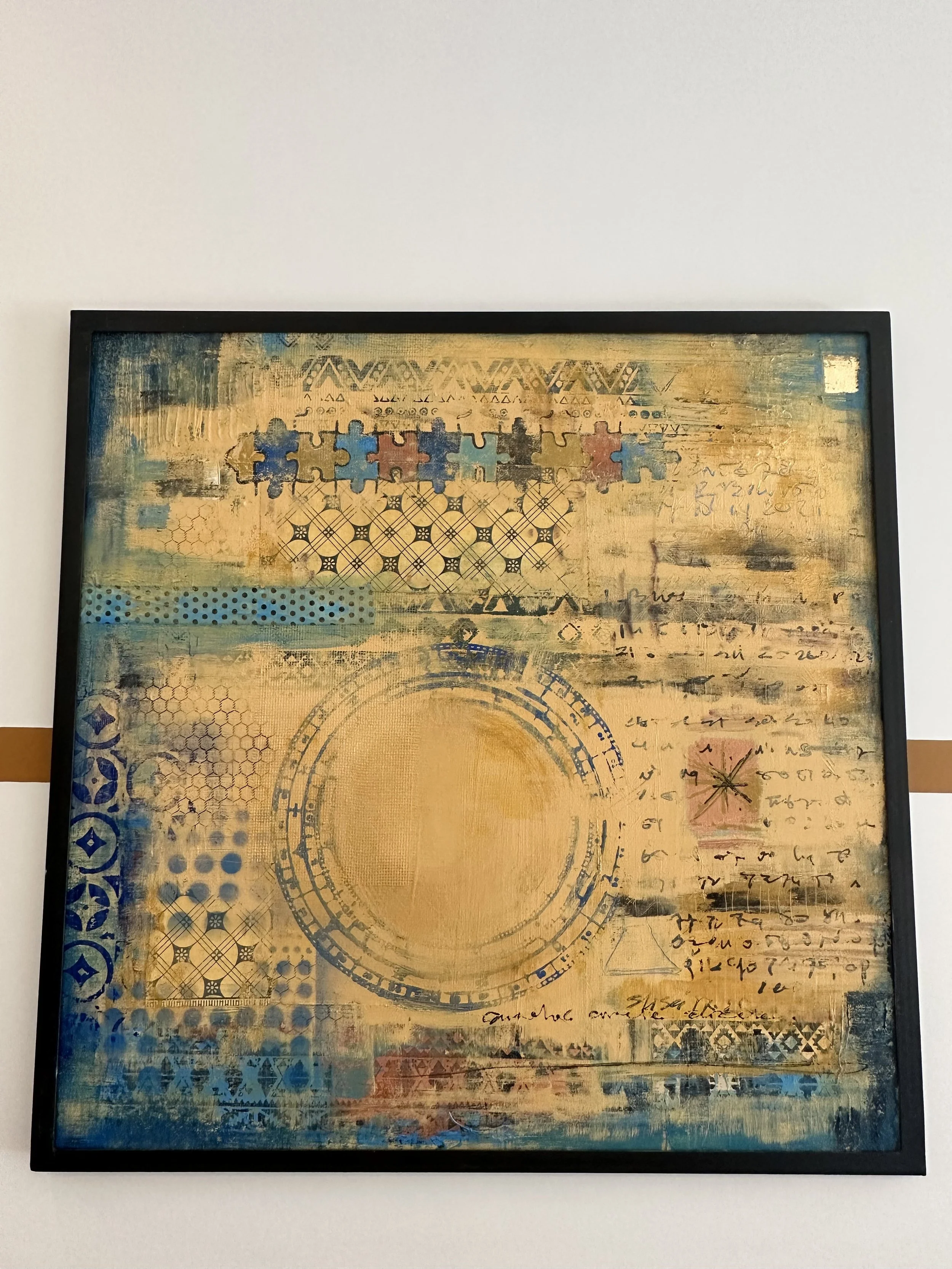 Abstract mixed media artwork featuring geometric patterns, handwritten script, puzzle pieces, and a large central circle, primarily in shades of gold, blue, black, and red, framed and hung on a white wall.