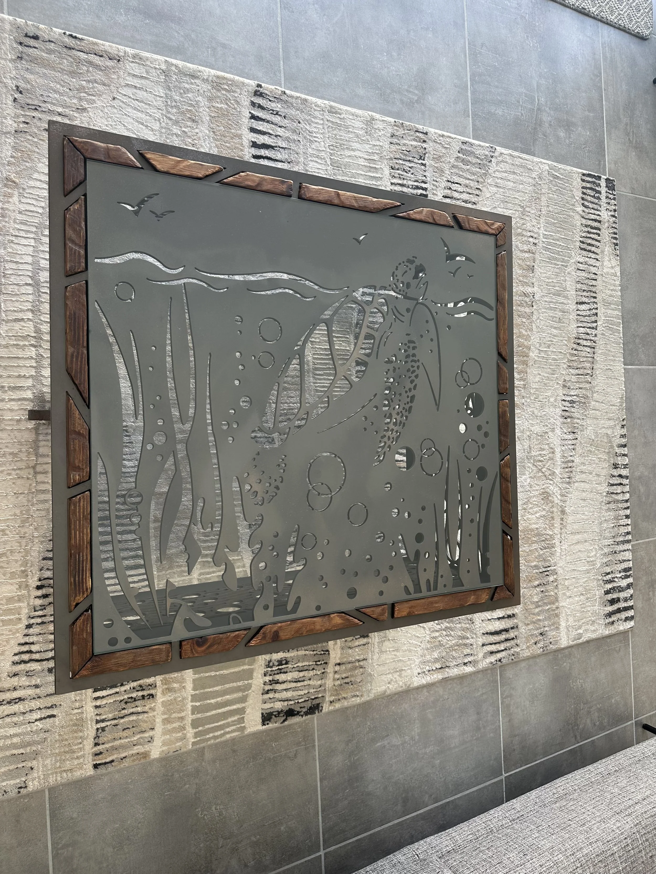 Decorative metal panel featuring a fish, seaweed, and bubbles, framed with wood, placed on a textured beige and gray area rug on a gray tiled floor.