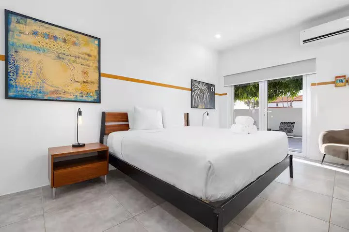 Modern hotel room with a large white bed, wooden nightstand with a lamp, colorful artwork on the wall, sliding glass door leading to a balcony with outdoor seating, and white walls with orange stripe detail.