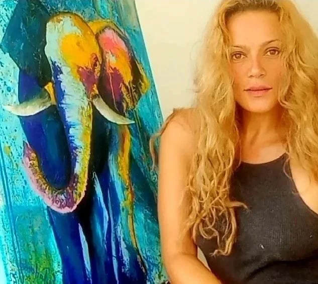 A woman with long curly blonde hair standing next to a colorful abstract painting of an elephant.
