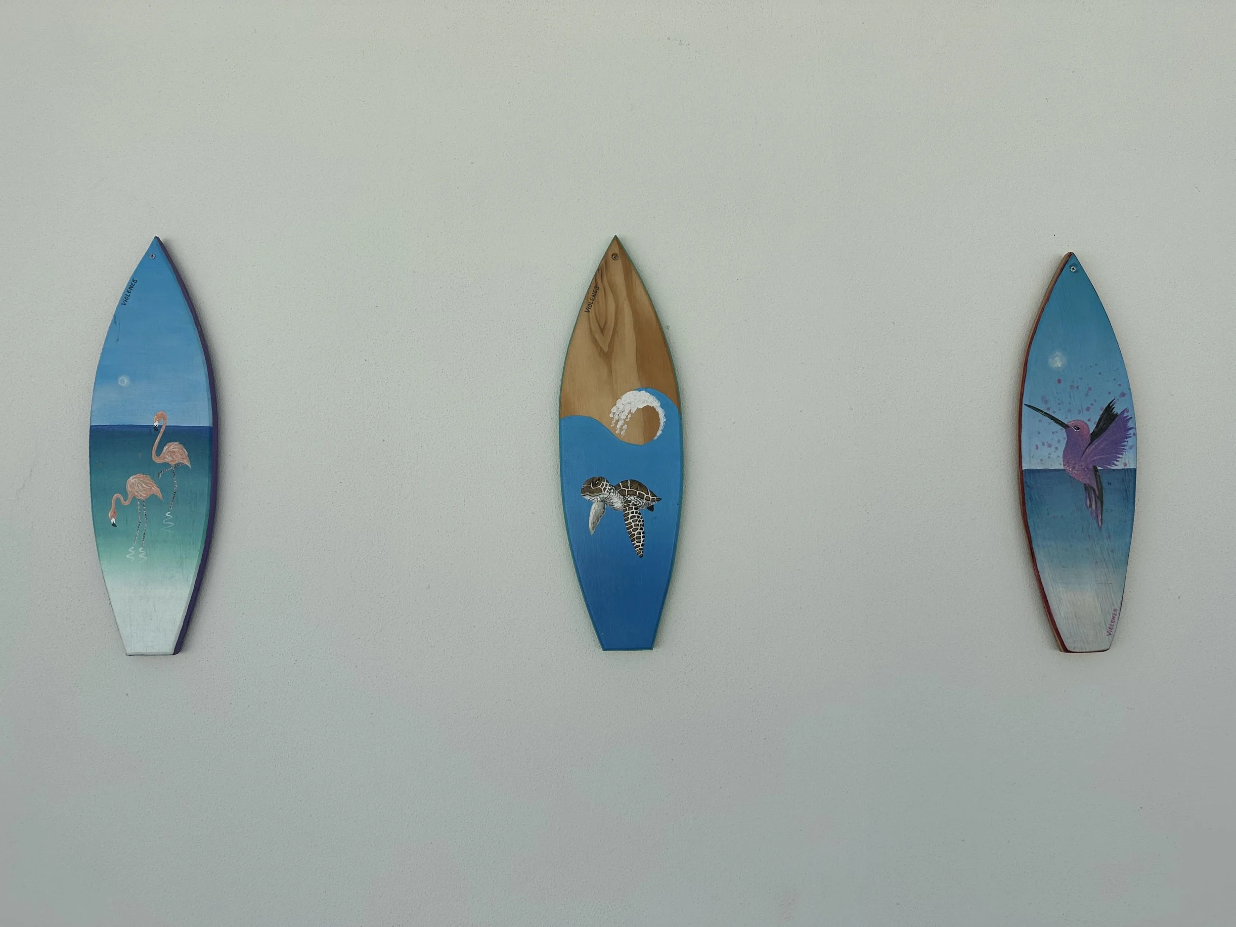 Three decorative paddle-shaped art pieces hanging on a wall, each depicting beach and ocean scenes with flamingos, a turtle, and a hummingbird.