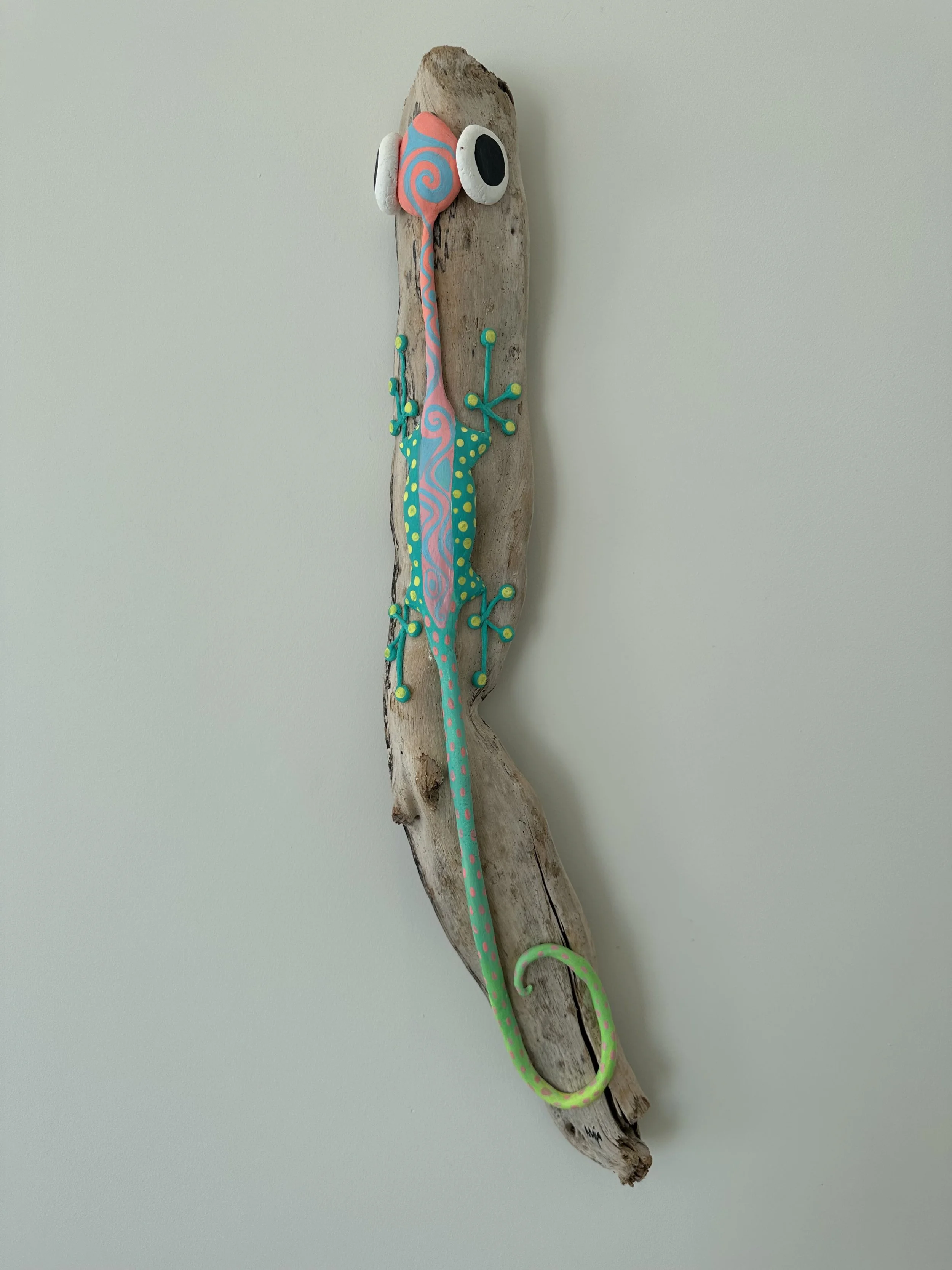 Colorful gecko sculpture with pink, blue, green, and black accents mounted on a piece of weathered wood on a white wall.