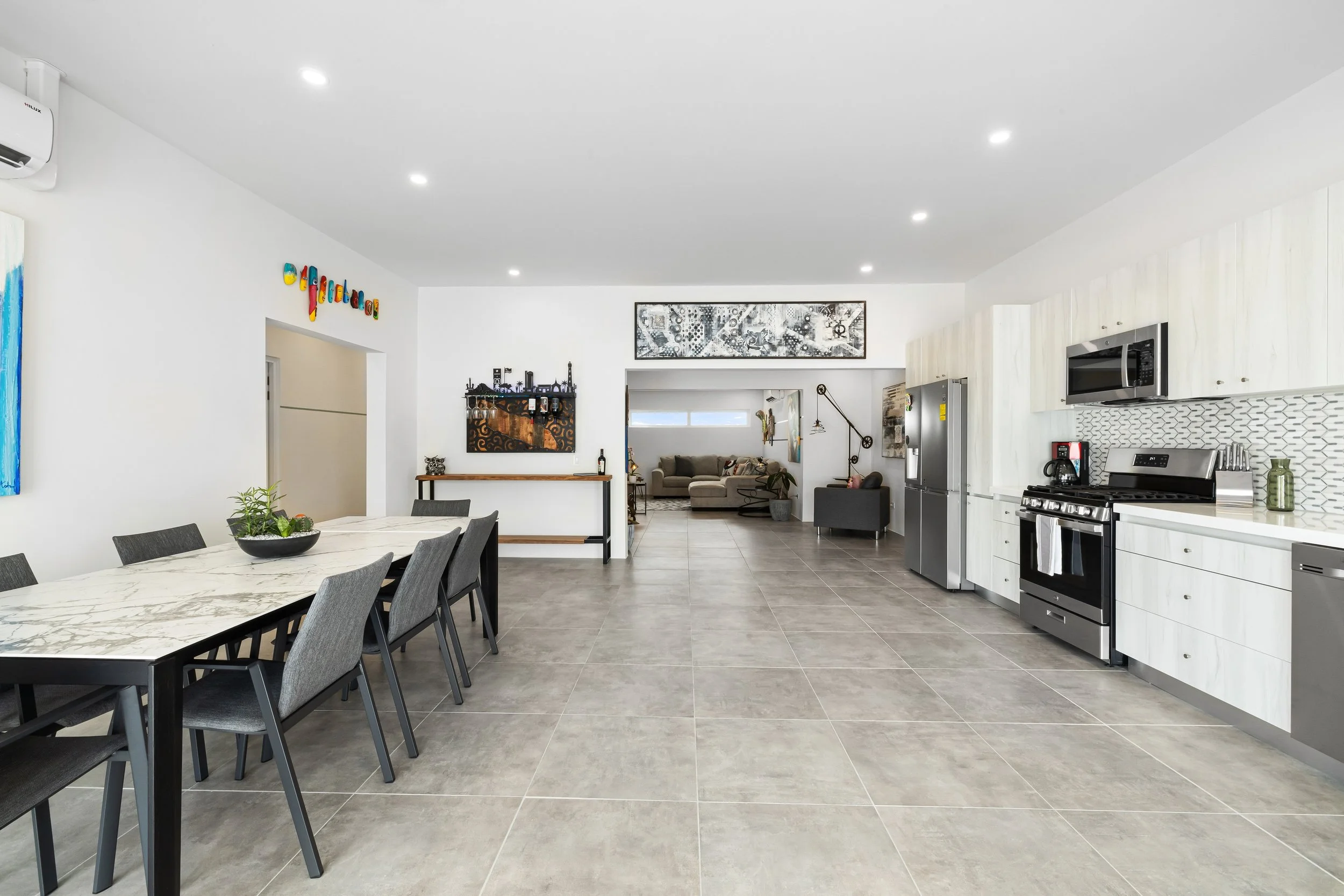 Spacious open-concept kitchen and dining area with modern appliances, a marble dining table with eight chairs, and a view into a living room with sofas and wall art.
