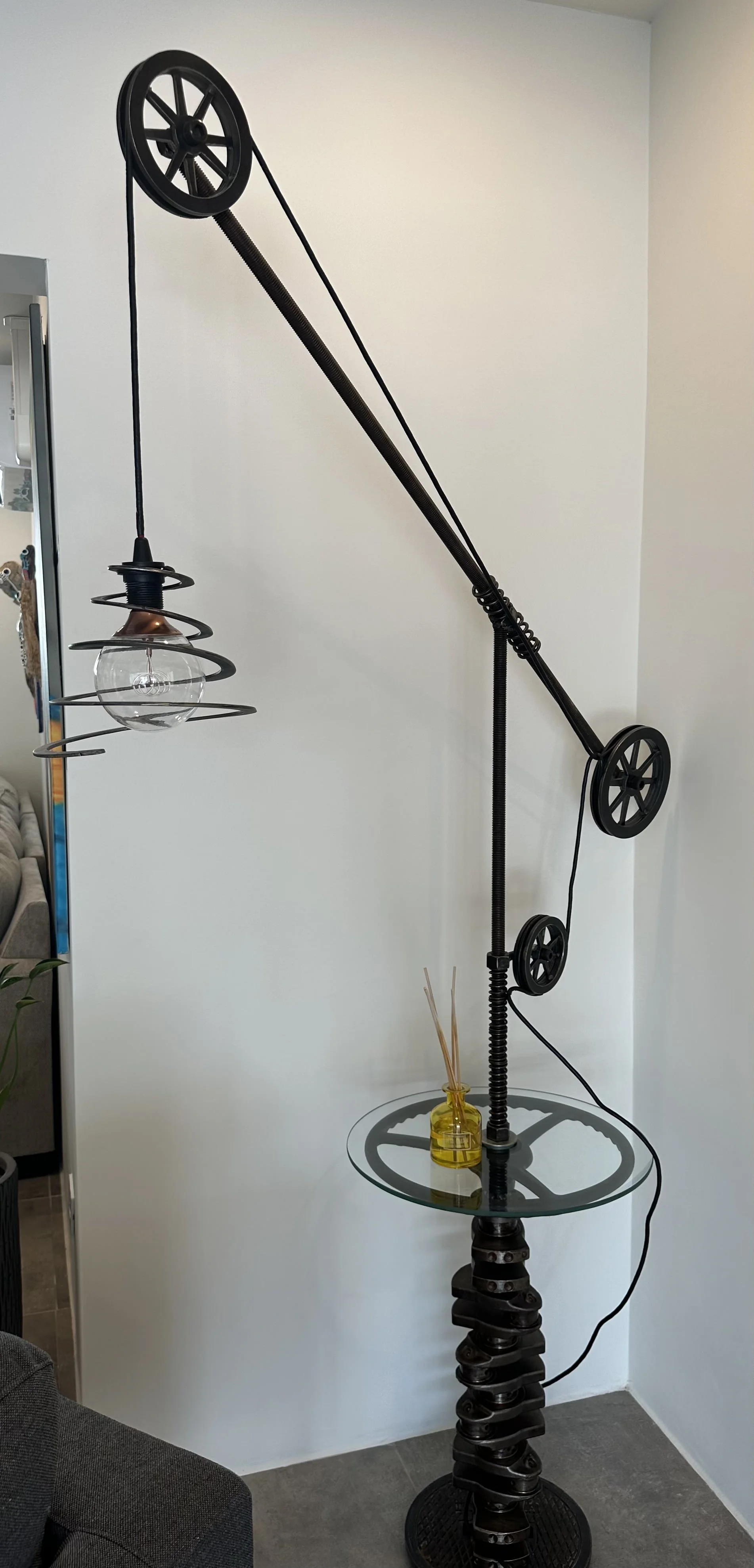 Unique floor lamp with a metal coil base, glass tabletop, and a long adjustable arm with pulleys, decorative lightbulb, and a yellow bottle with reed diffusers.
