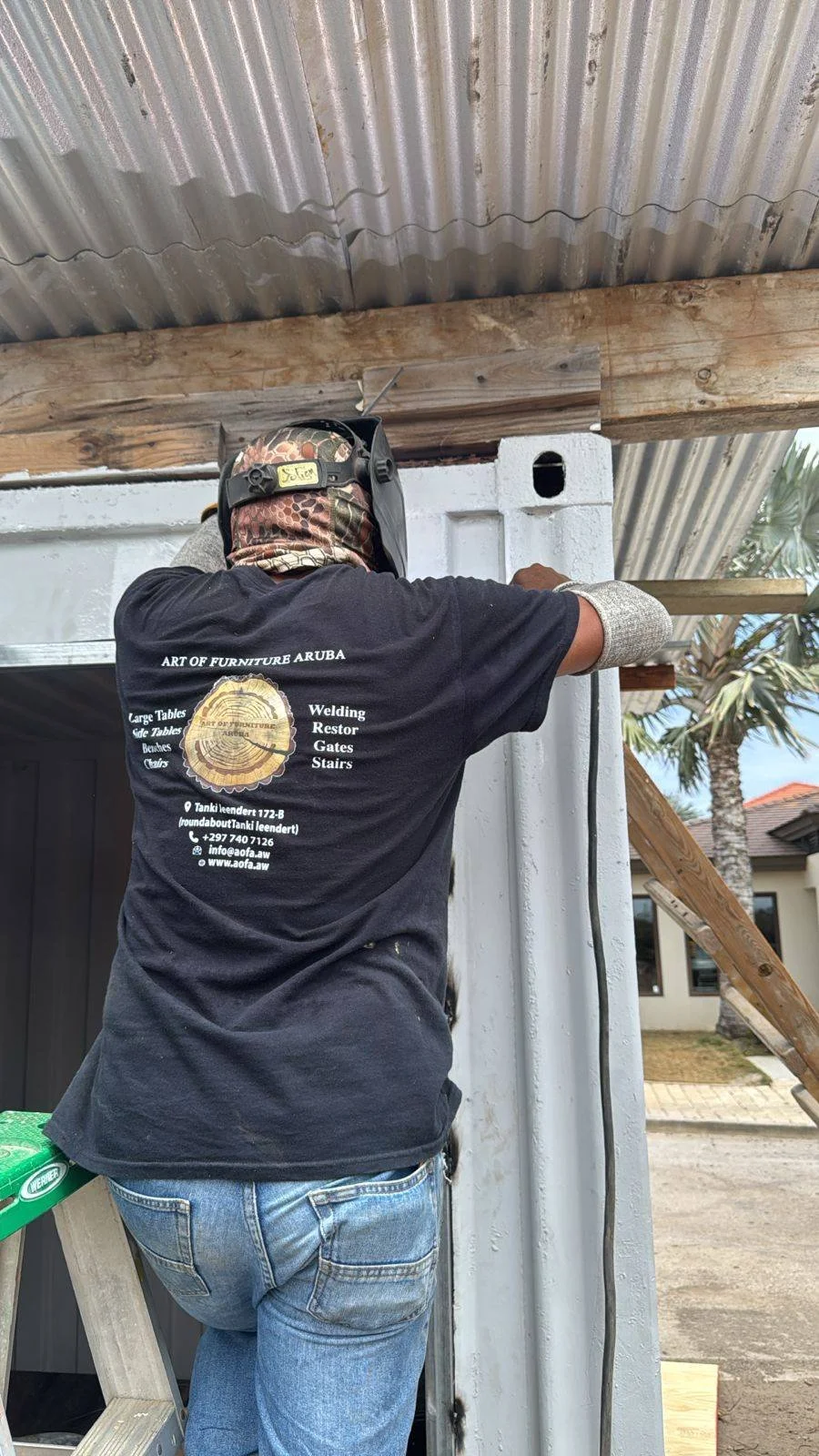A person wearing a welding helmet and gloves working on a construction project, attaching a wooden beam to a concrete or metal wall under a corrugated metal roof.