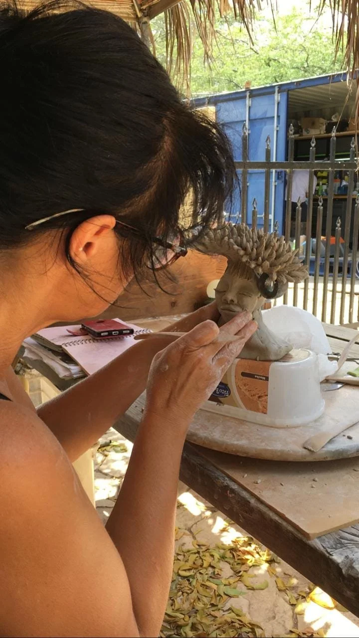 A woman is sculpting a ceramic or clay bust of a person with a decorative headdress, working outdoors under a thatched roof with trees and a blue fencing in the background.