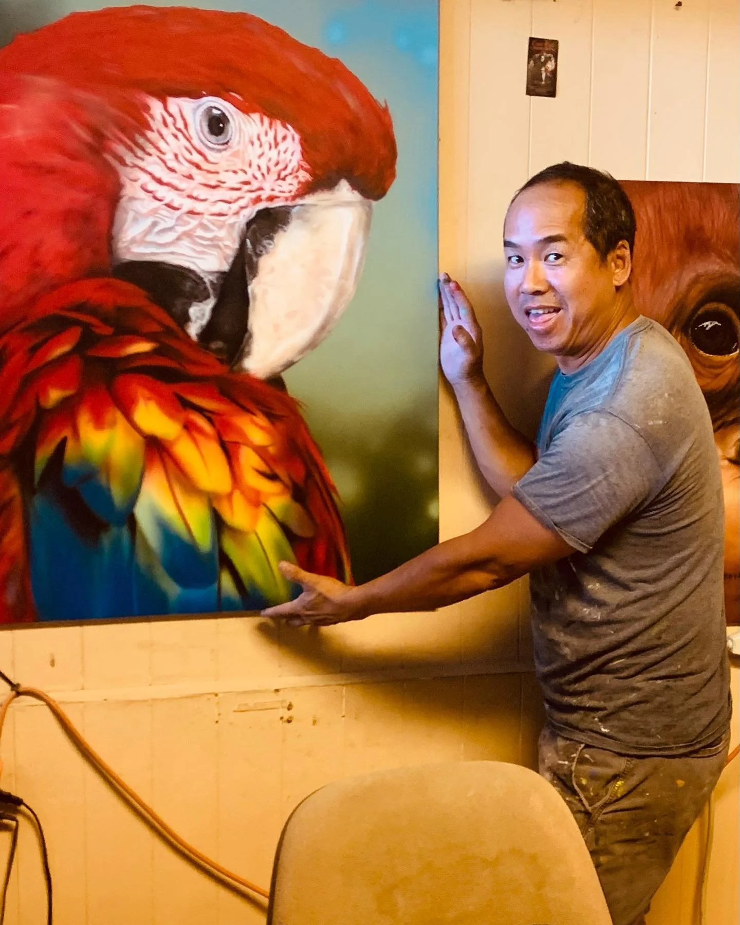 A man is posing with a large painting of a colorful parrot, and he is touching the painting with both hands and smiling.
