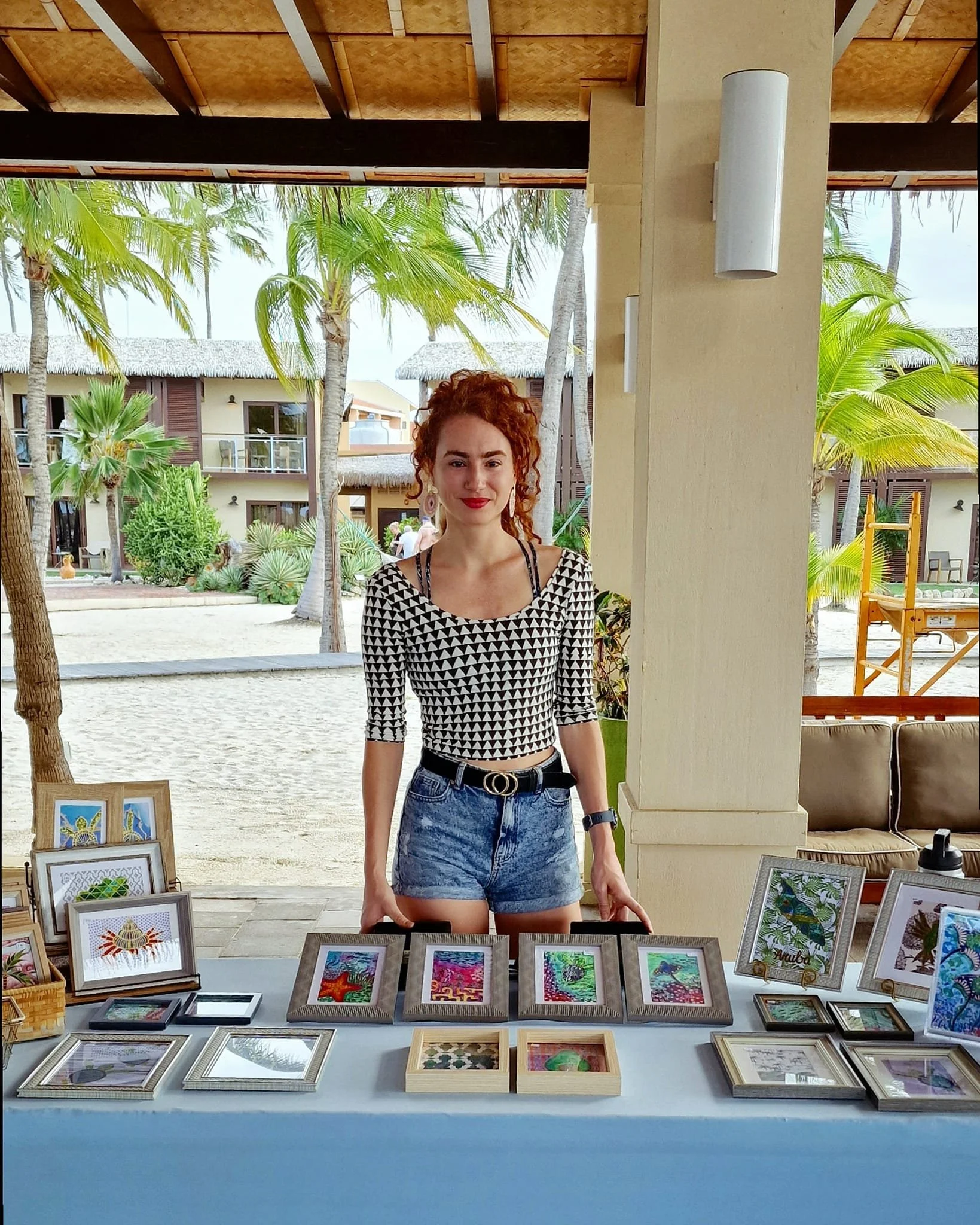 A woman with curly red hair and wearing a black and white patterned top stands behind a table displaying framed artwork at an outdoor market near a beach with palm trees and buildings in the background.