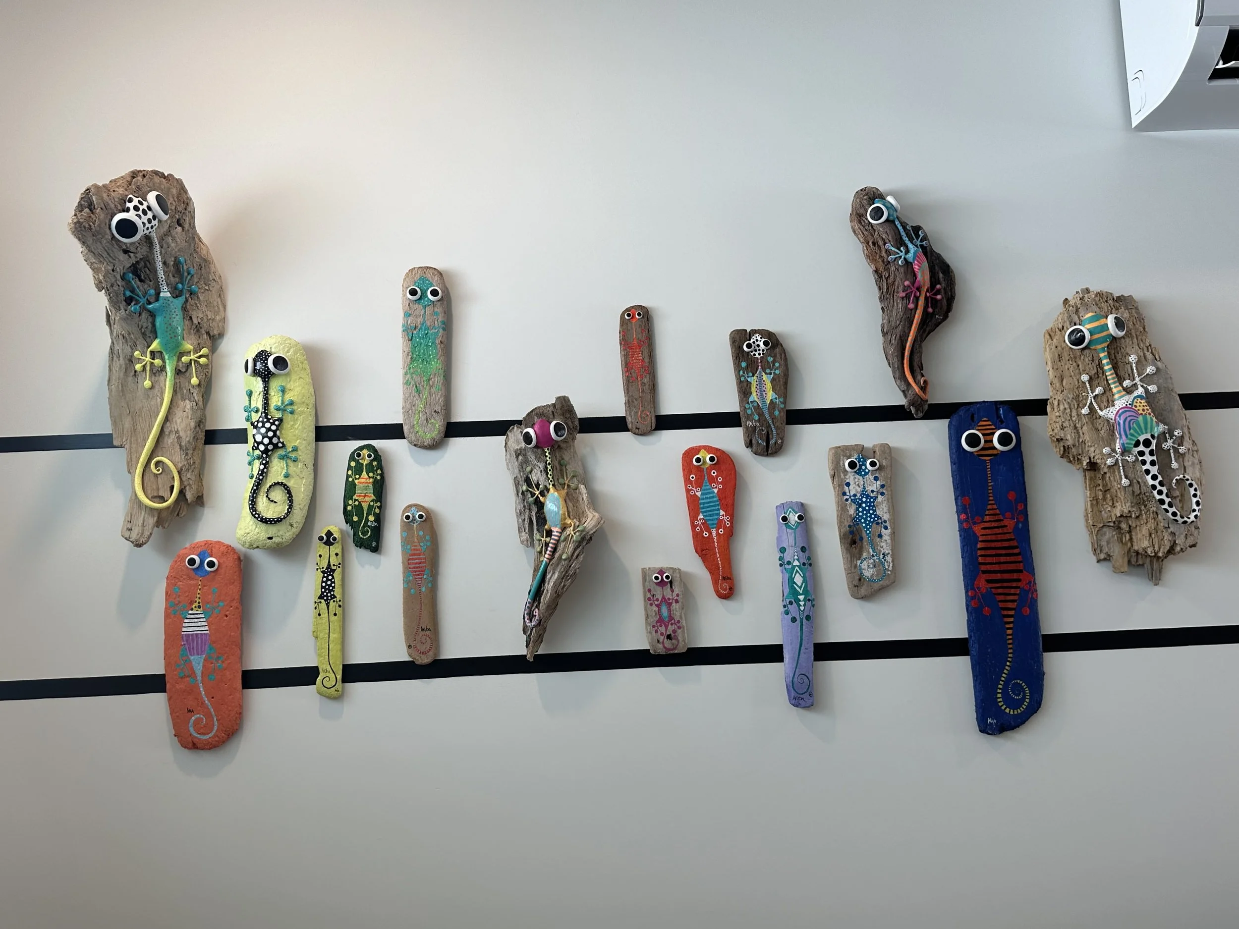 Colorful handcrafted gecko and salamander decorations attached to wooden pieces, with googly eyes, on a white wall with black lines.