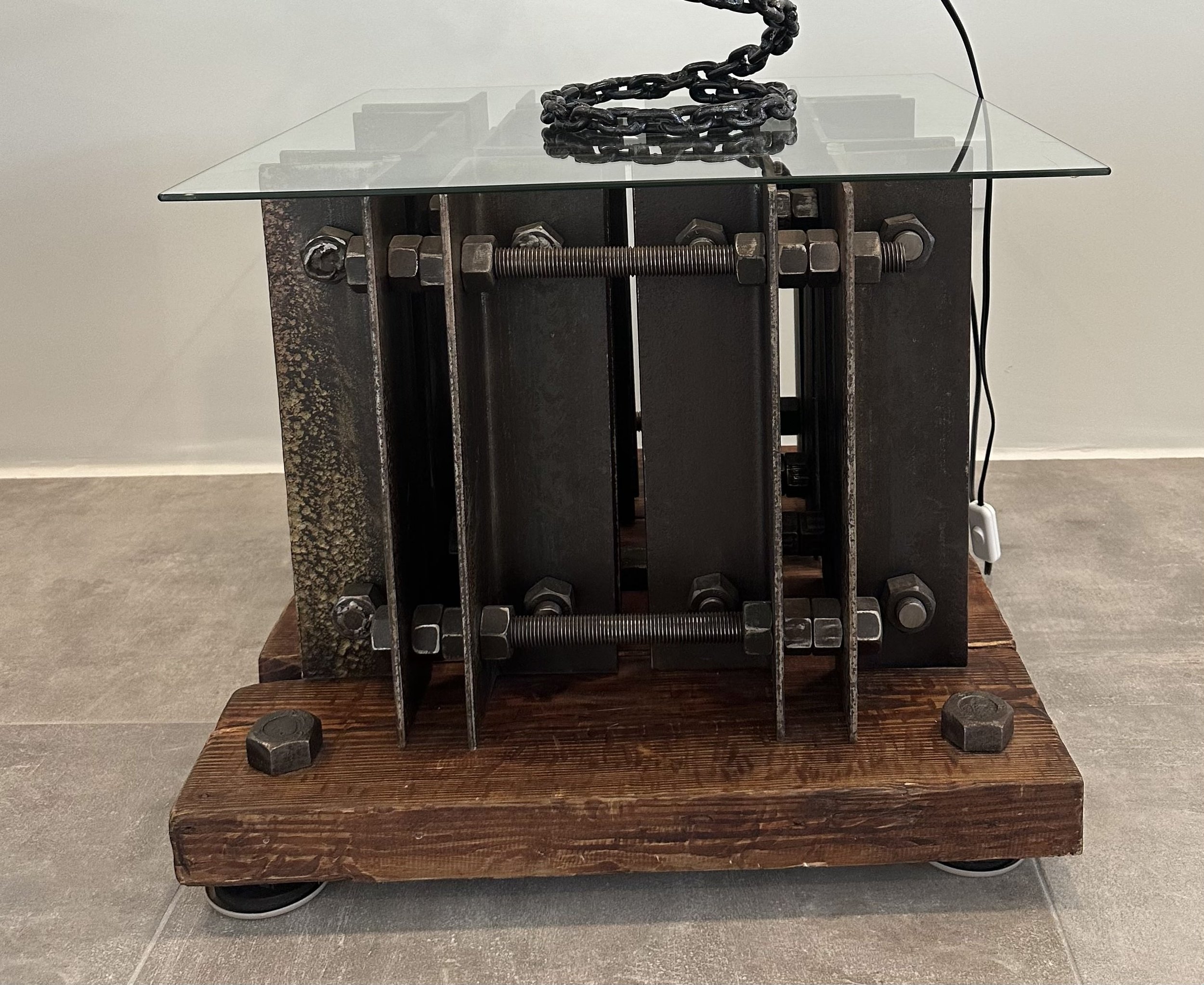 An art sculpture made of metal and wood with a glass top, placed on a wooden base with wheels. The sculpture resembles a machine, with large nuts and bolts, metal plates, and a chain on top.