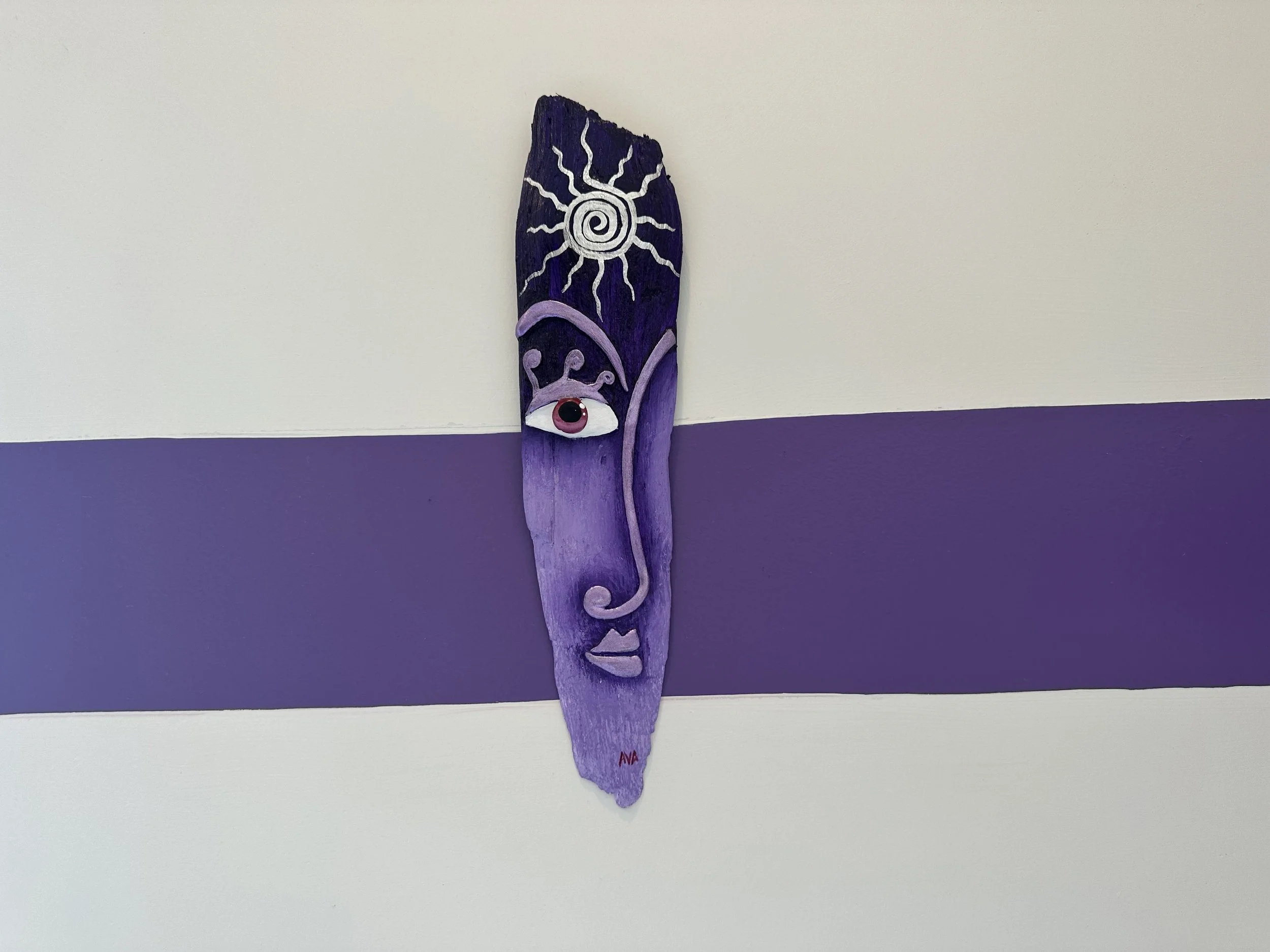 Decorative purple wooden panel with a painted face featuring an eye and lips, and a sun with rays painted above, installed on a white wall with a purple stripe.