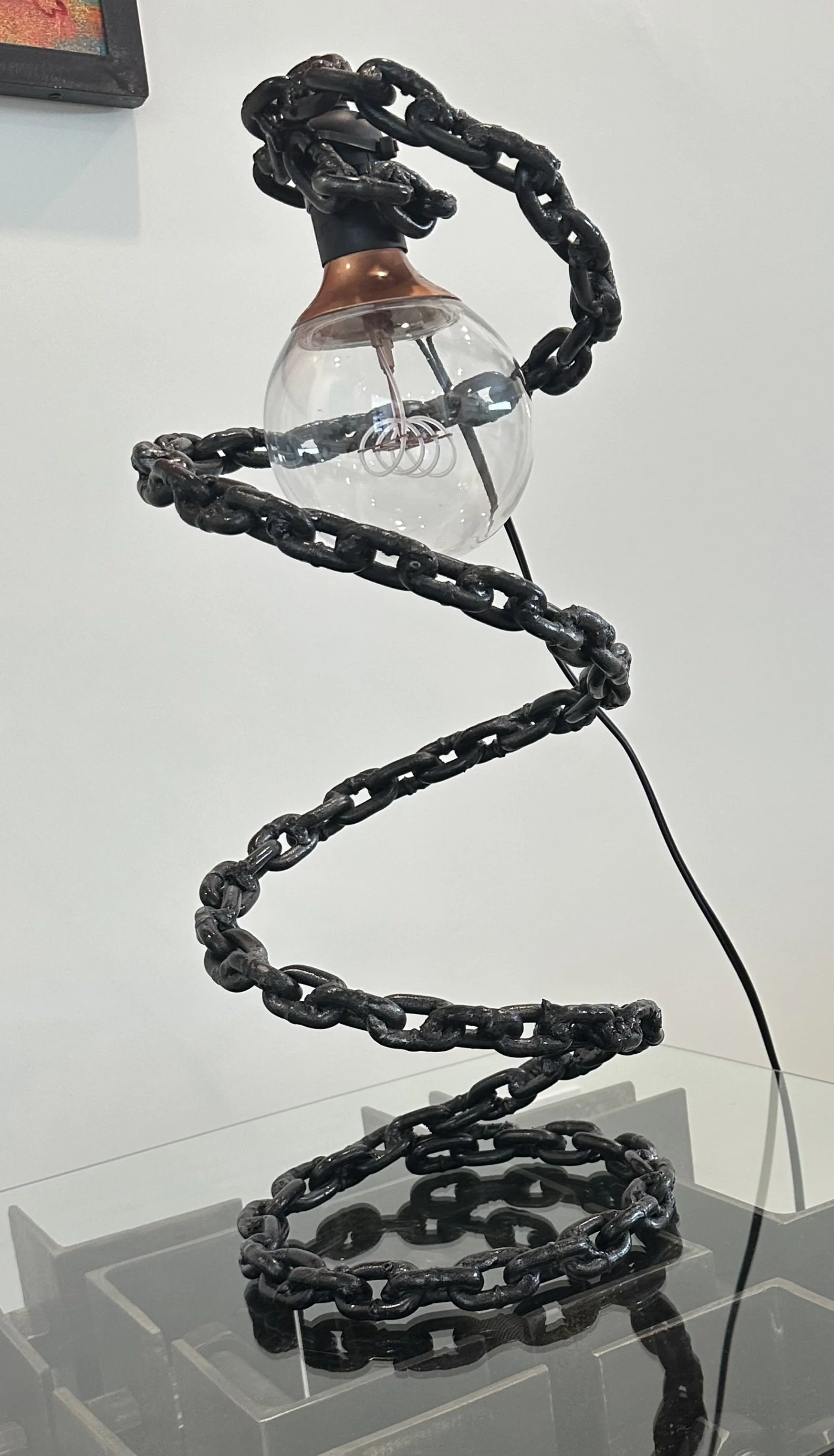 A decorative sculpture resembling a DNA helix, made of black metal chains, with a light bulb in the middle, placed on a glass surface.