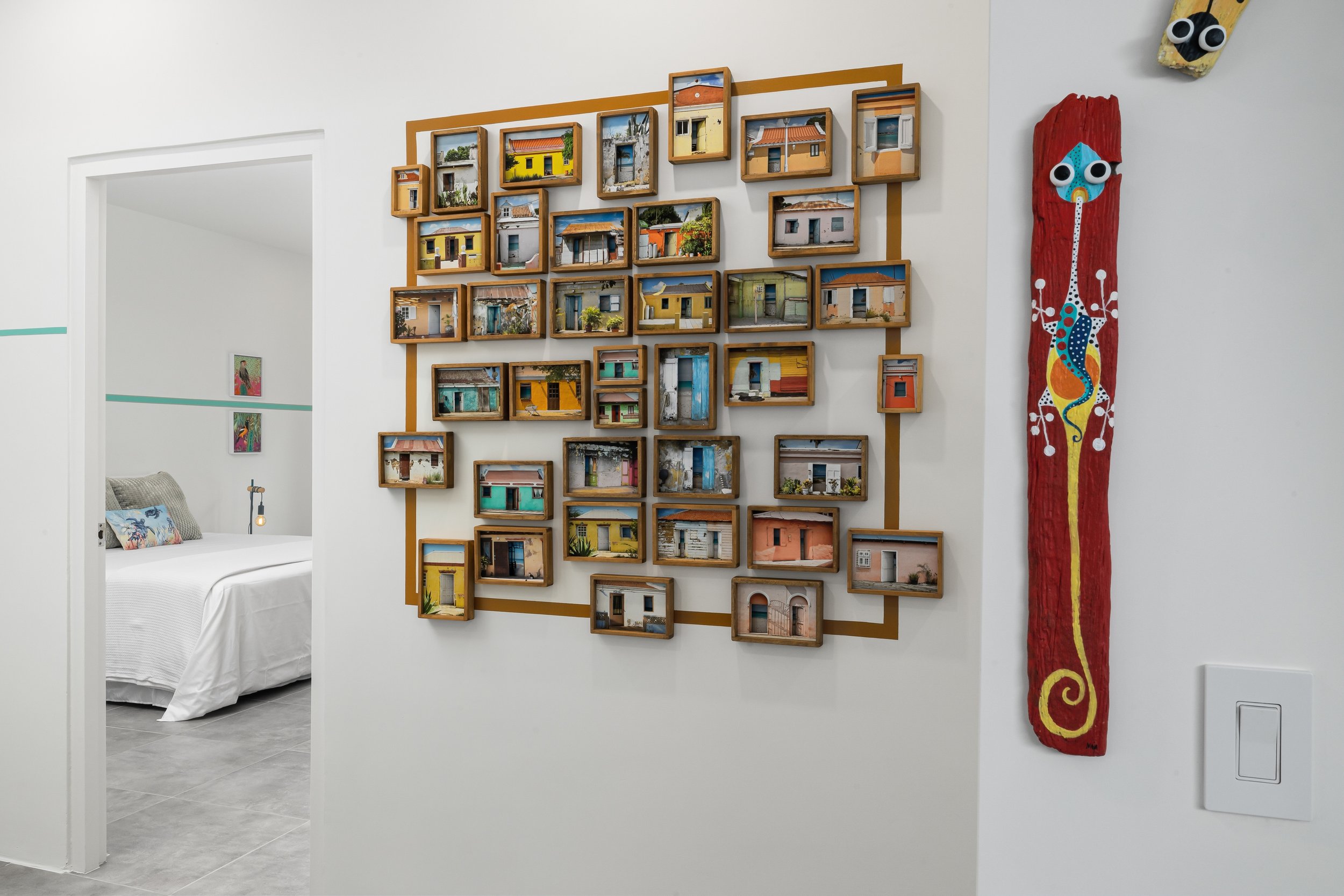 Wall art display of multiple framed photographs of colorful houses, arranged in a grid pattern with a wooden border, next to a vertically elongated painted wooden piece with a stylized animal face, in a bright and modern interior.