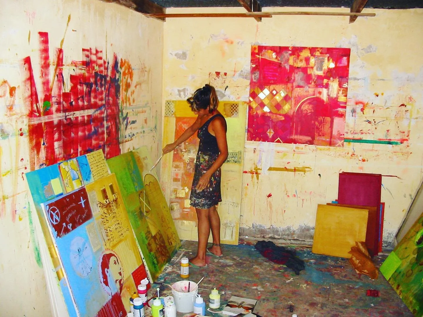 A woman painting and working on multiple colorful paintings in an art studio with a yellowed, stained wall and various painting supplies on the floor.