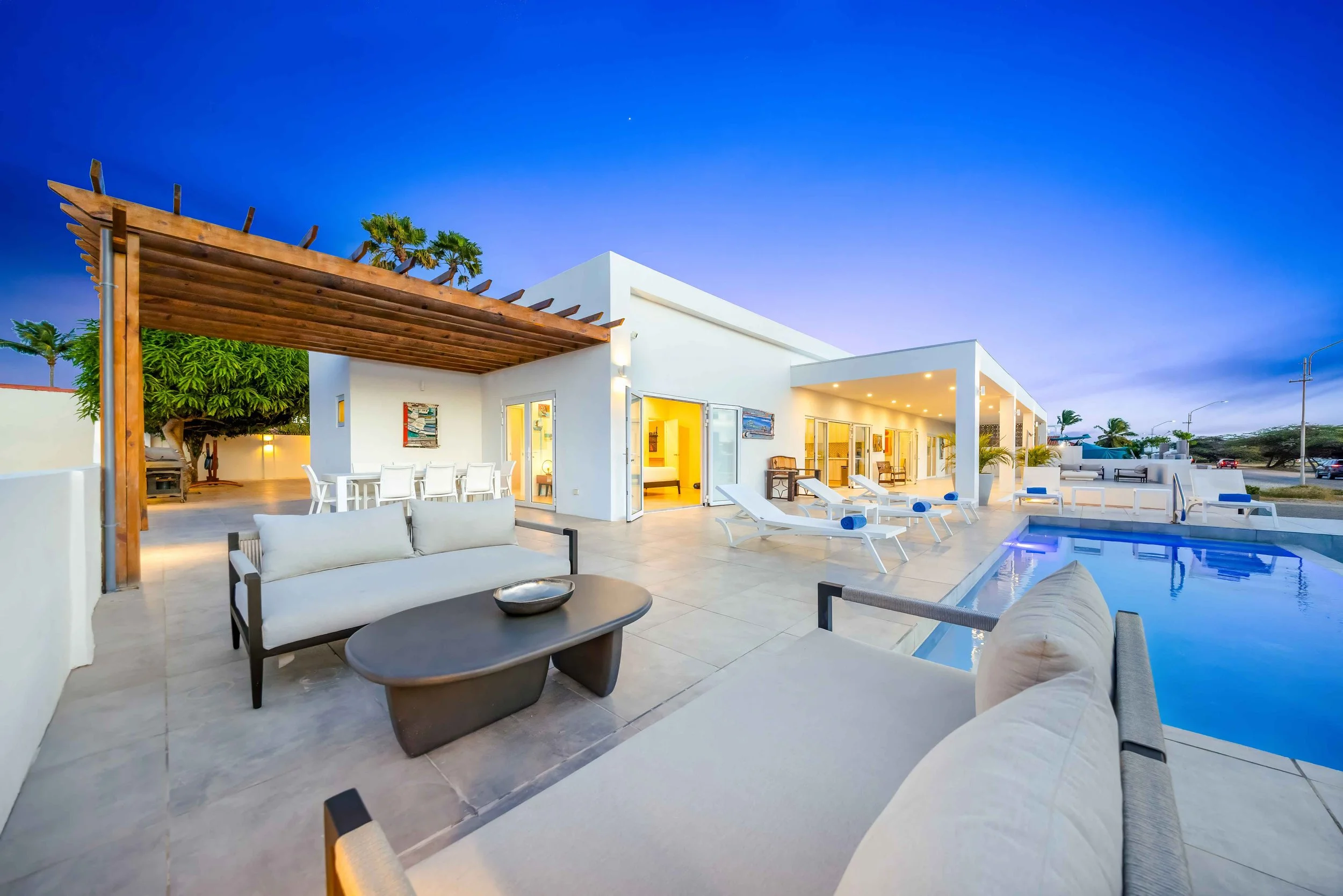Modern white house with outdoor patio, swimming pool, and lounge chairs during evening. Wooden pergola over dining area, bright lighting inside and outside, surrounded by minimal decor and palm trees.