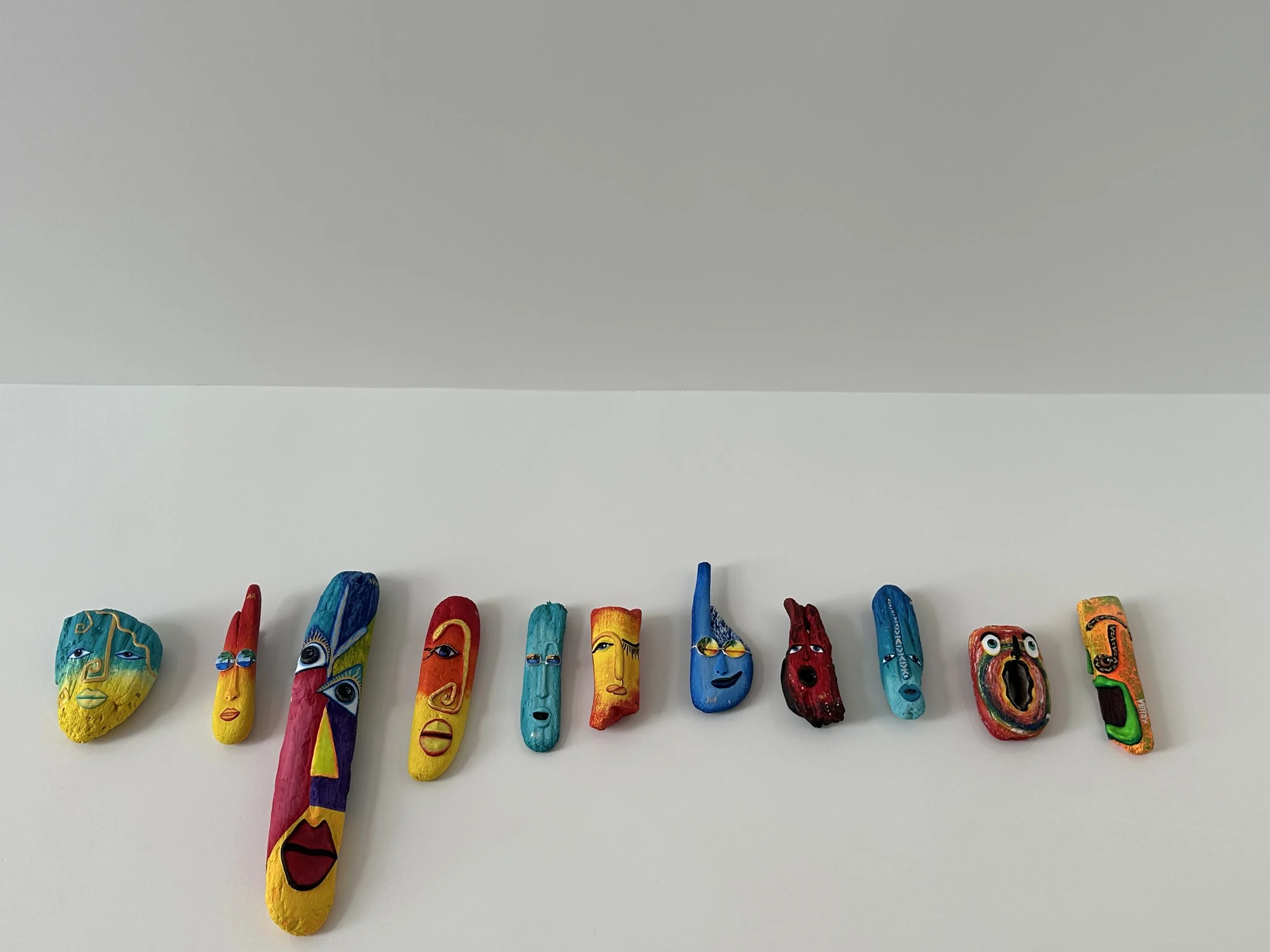 Colorful painted rocks with abstract faces and unique designs are arranged in a line on a white surface.