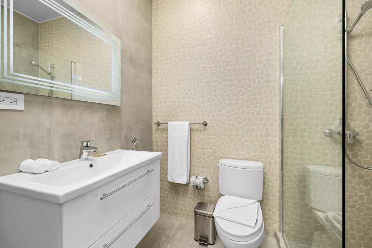 Modern bathroom with beige textured wall tiles, a white sink with a large mirror above, a toilet with a folded towel on the lid, a towel bar with a white towel, a small trash can, and a glass shower enclosure with a handheld showerhead.