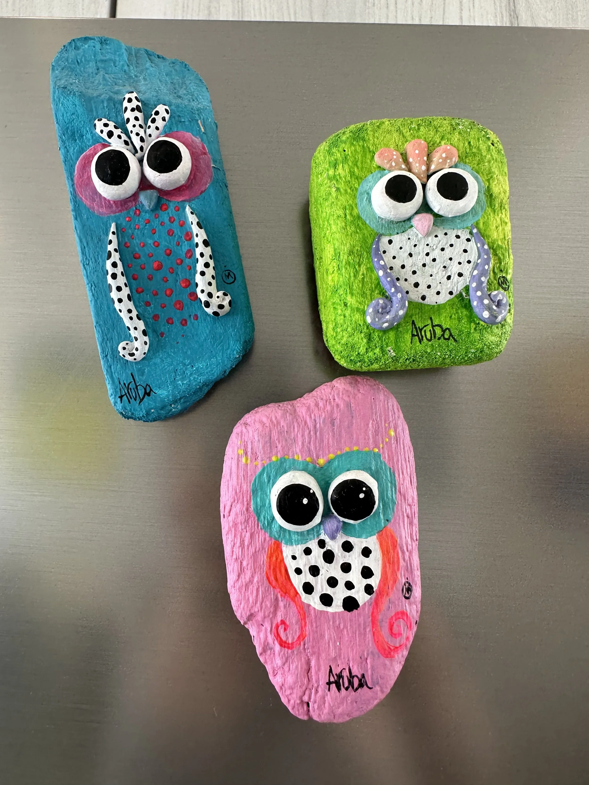 Three painted rocks with whimsical owl designs on a metallic surface.