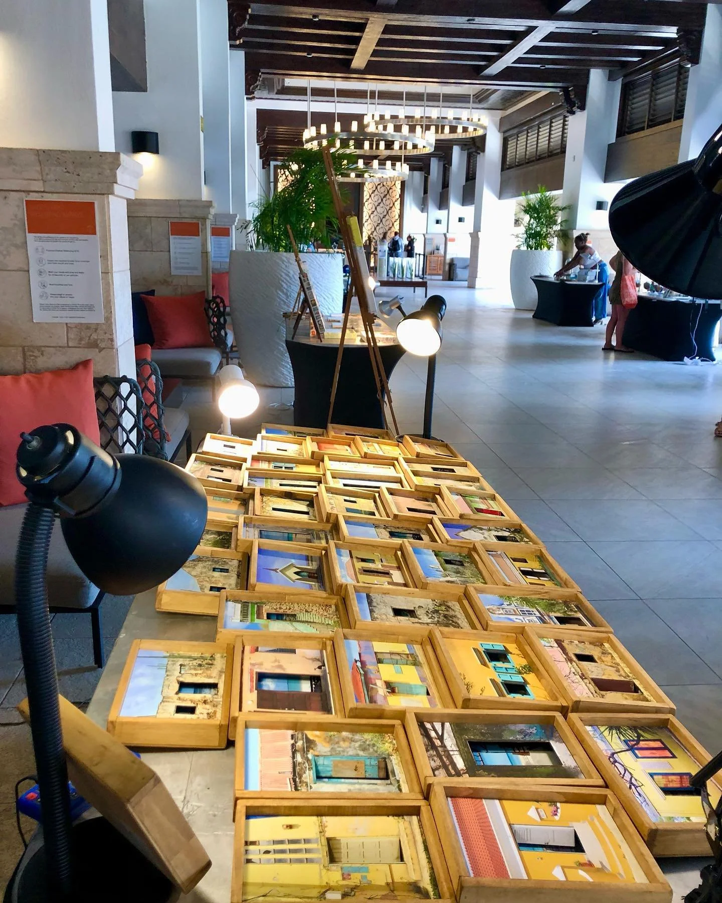A table with numerous framed photographs or artwork displayed, set in a spacious, modern interior with high ceilings, large plants, and people in the background, possibly at an art show or gallery.