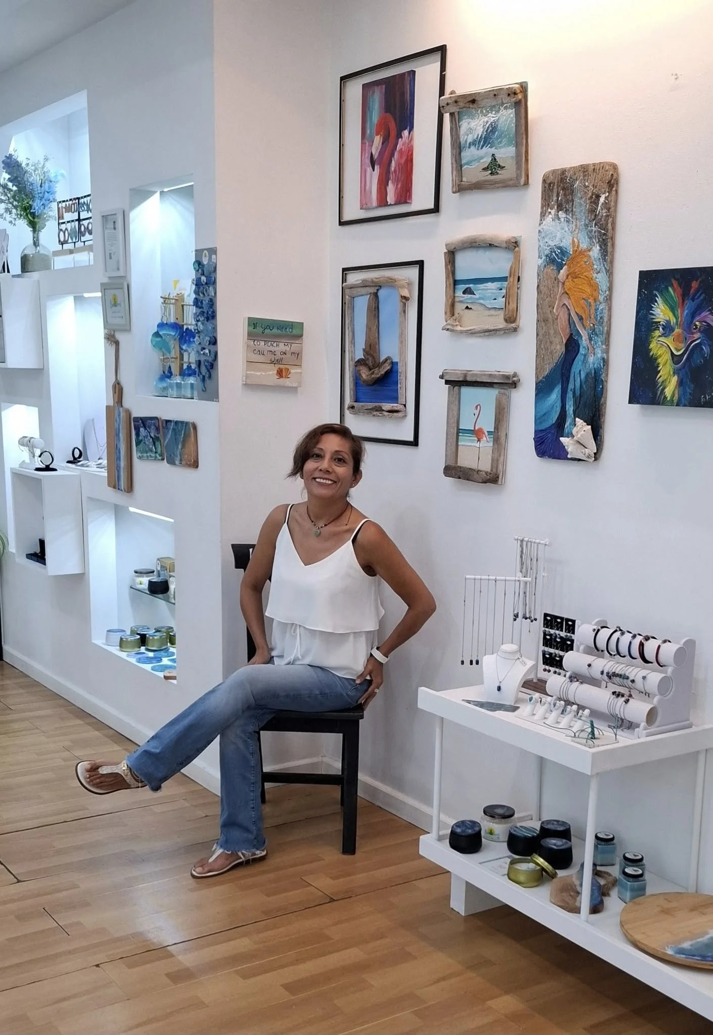A woman sitting on a black chair inside an art gallery or boutique, smiling at the camera. The wall behind her displays various coastal-themed paintings and small art pieces, with shelves holding jewelry and decorative items. The flooring is wooden.