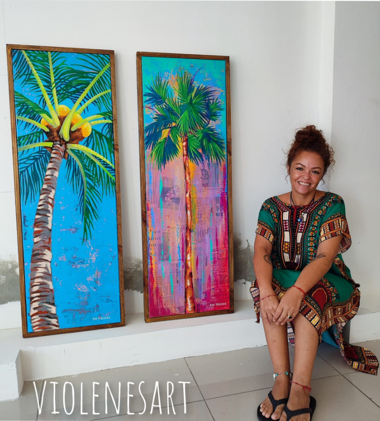 Woman with curly hair smiling, sitting on the floor next to two colorful paintings of palm trees, one with a blue background and the other with a pink and purple background, displayed on a white wall.