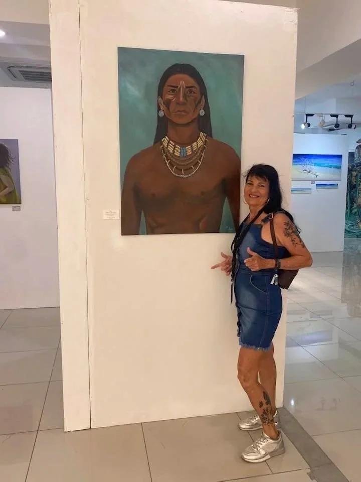 A woman with a tattoo on her leg and arm, wearing a denim dress and sneakers, stands next to a large portrait of a topless Indigenous woman adorned with jewelry, in an art gallery.