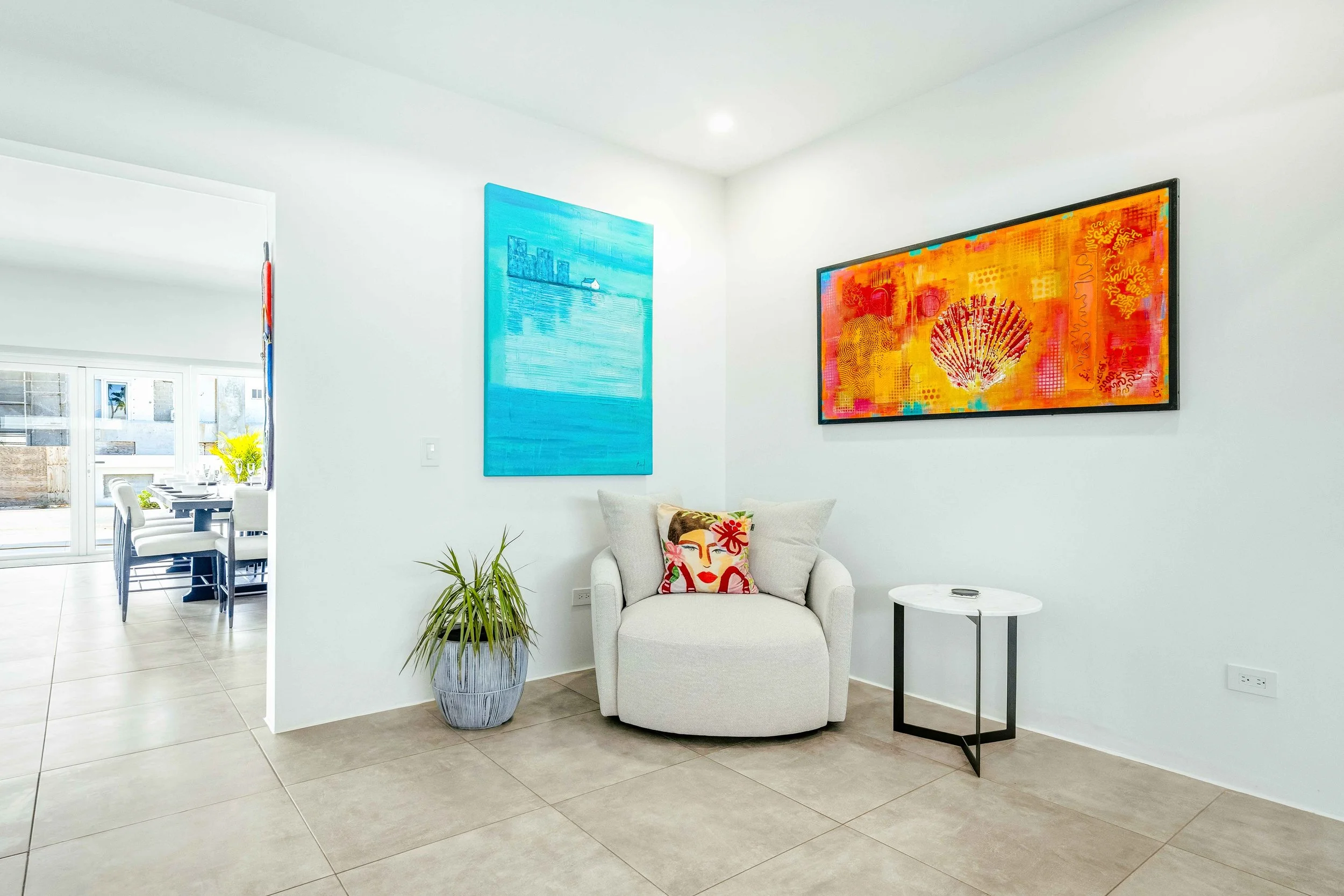 Living room corner with a white armchair, colorful cushion, potted plant, two vibrant wall paintings, and a small round table.
