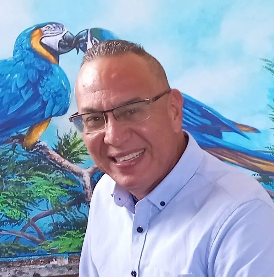 A smiling man wearing glasses in front of a large colorful painting of a blue-and-yellow macaw bird on a branch.