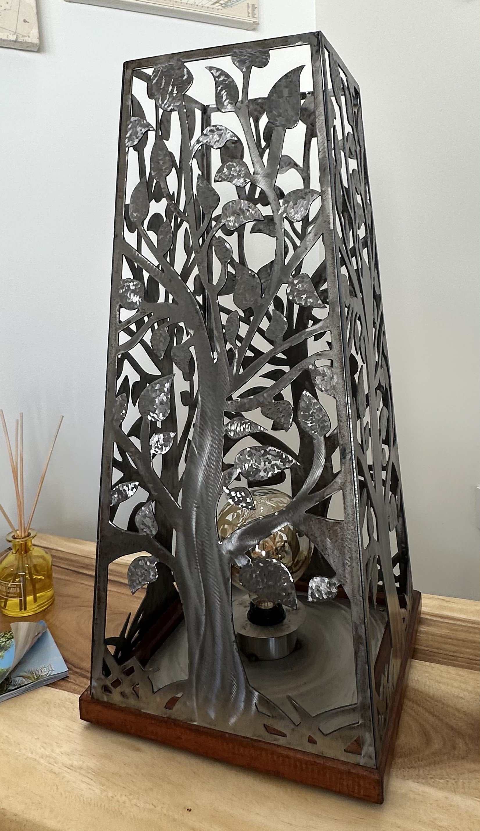 A decorative metal lamp with a tree and leaf design, placed on a wooden table alongside a yellow diffuser with reed sticks, a magazine, and a small glass container.
