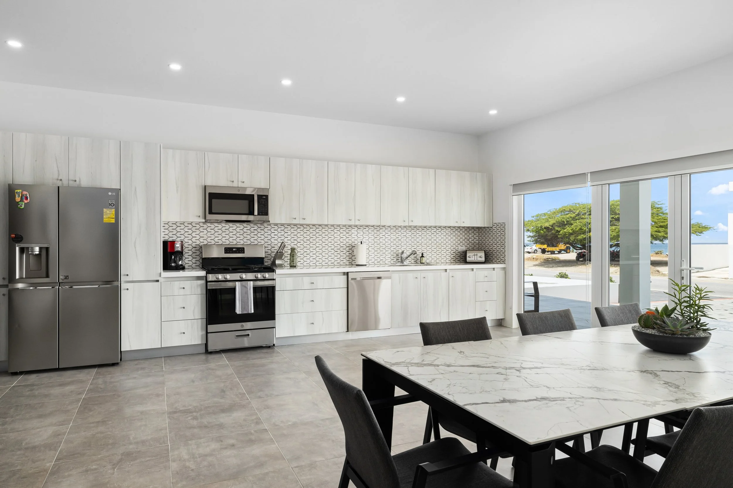 Modern kitchen with white cabinets, stainless steel appliances, and a large dining table with black chairs, overlooking an outdoor view through glass doors.