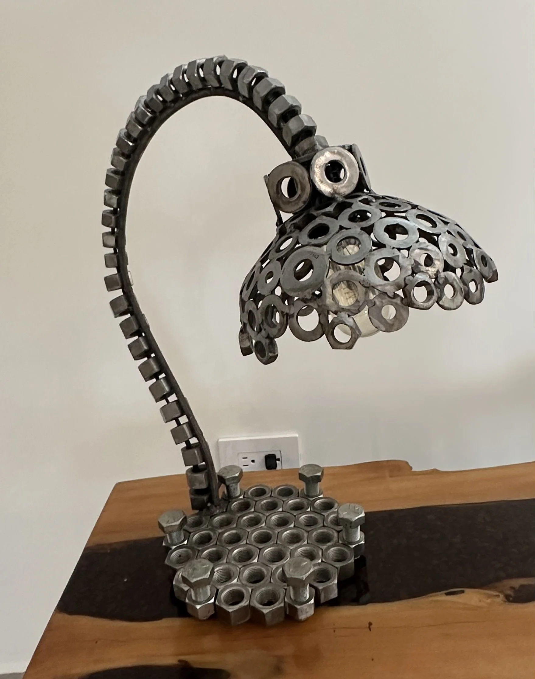 Lamp made of metallic nuts, bolts, and washers, designed to resemble an owl with a curved metal arm as the neck, mounted on a wooden surface.