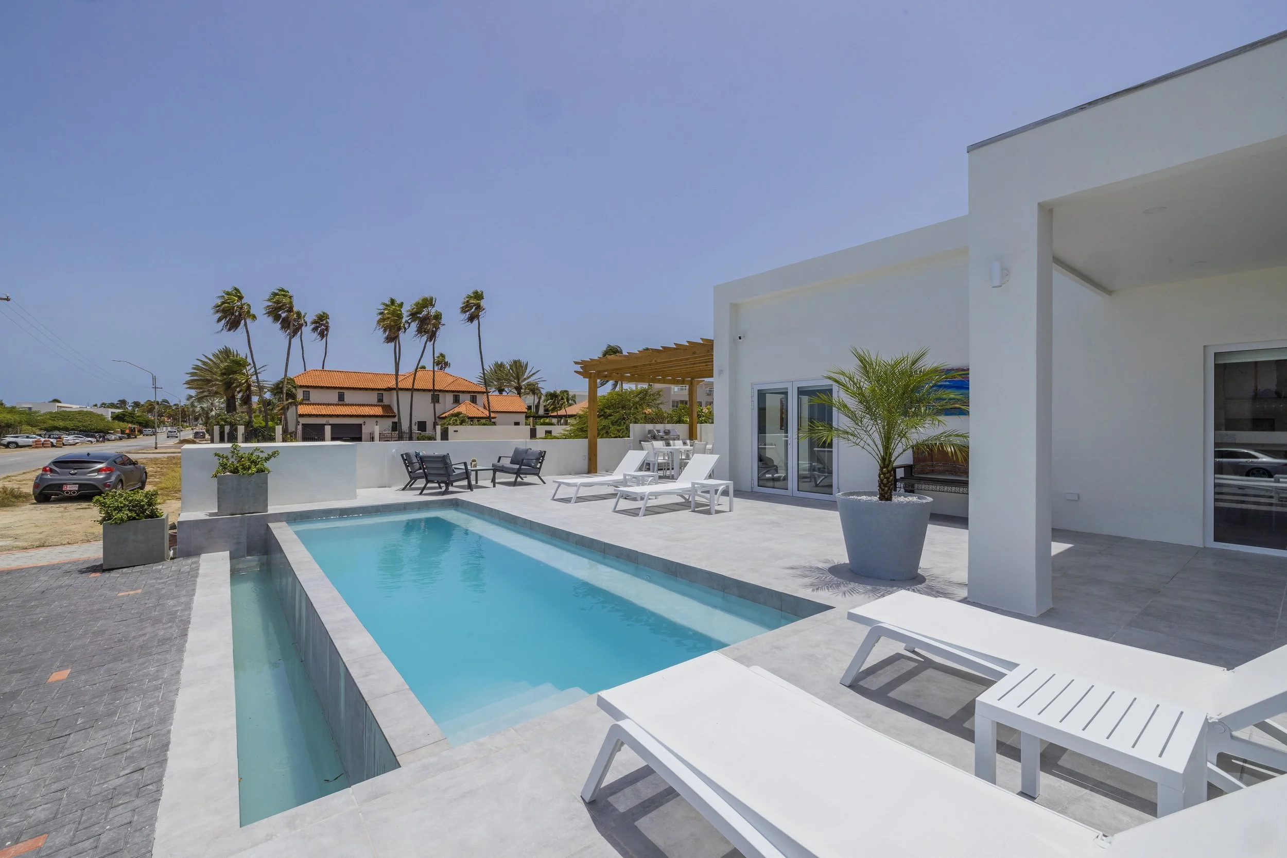 Modern house with a swimming pool, white patio furniture, potted plants, and a wooden pergola under a clear blue sky.