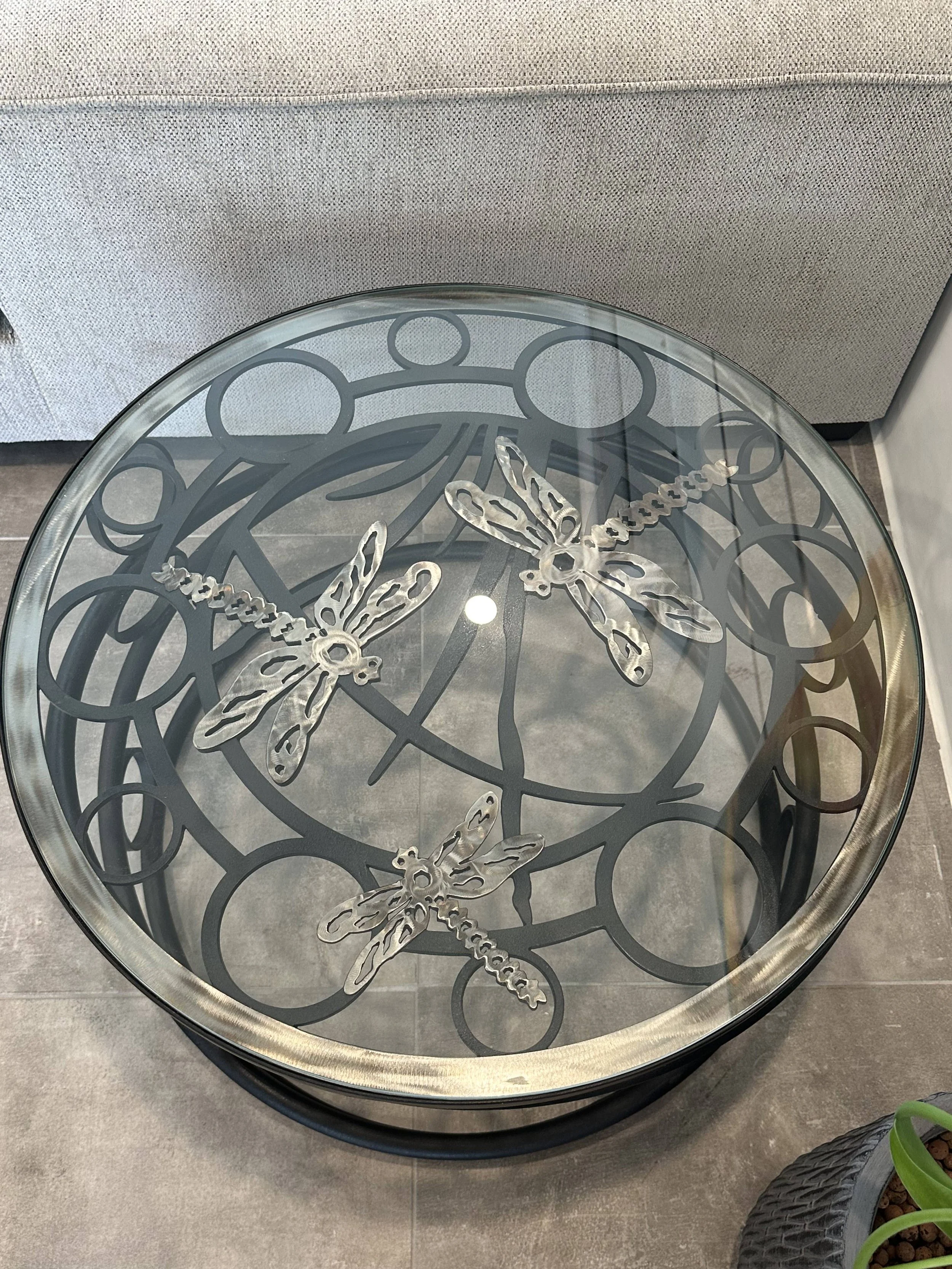 Round glass-top table with black metal base featuring decorative dragonfly designs, placed against a wall and next to a gray upholstered furniture piece.
