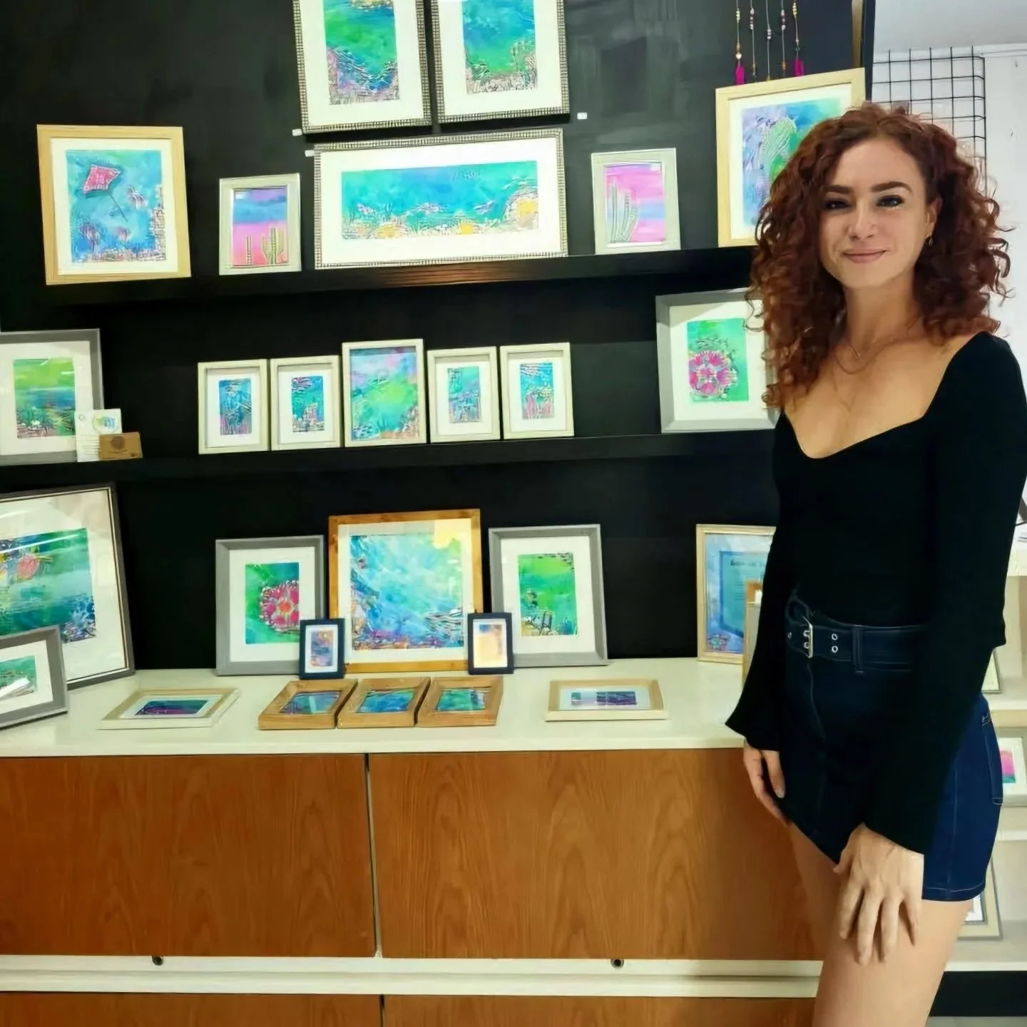 A woman with curly red hair wearing a black top and denim shorts standing next to a display of colorful framed artwork at an art gallery.