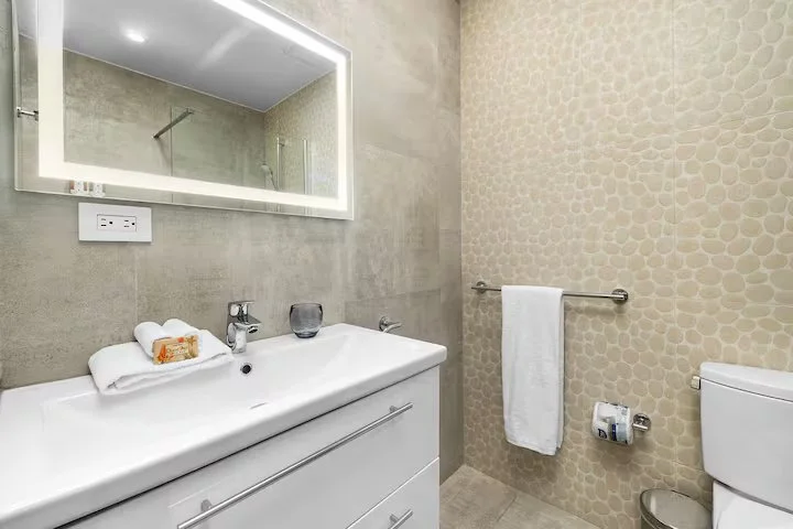 Modern bathroom with a white vanity, large mirror with backlit lighting, beige textured wall, towel rack with white towel, and toilet