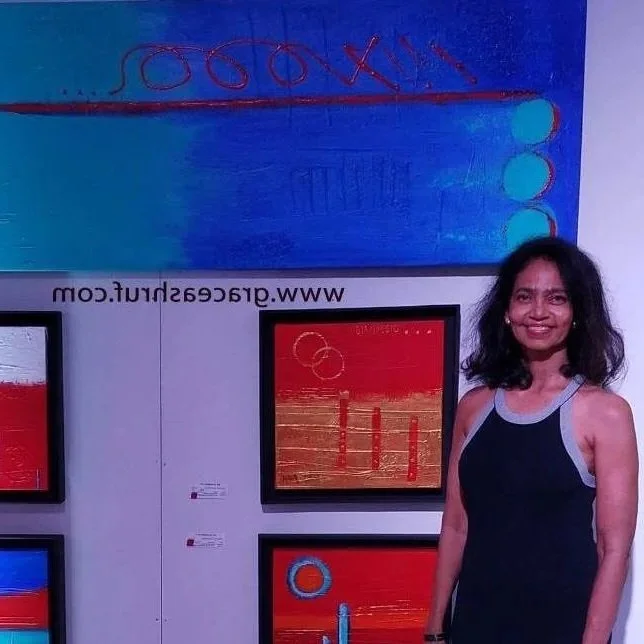 A woman with dark curly hair and a black and gray sleeveless top smiling while standing next to framed digital art on a gallery wall.