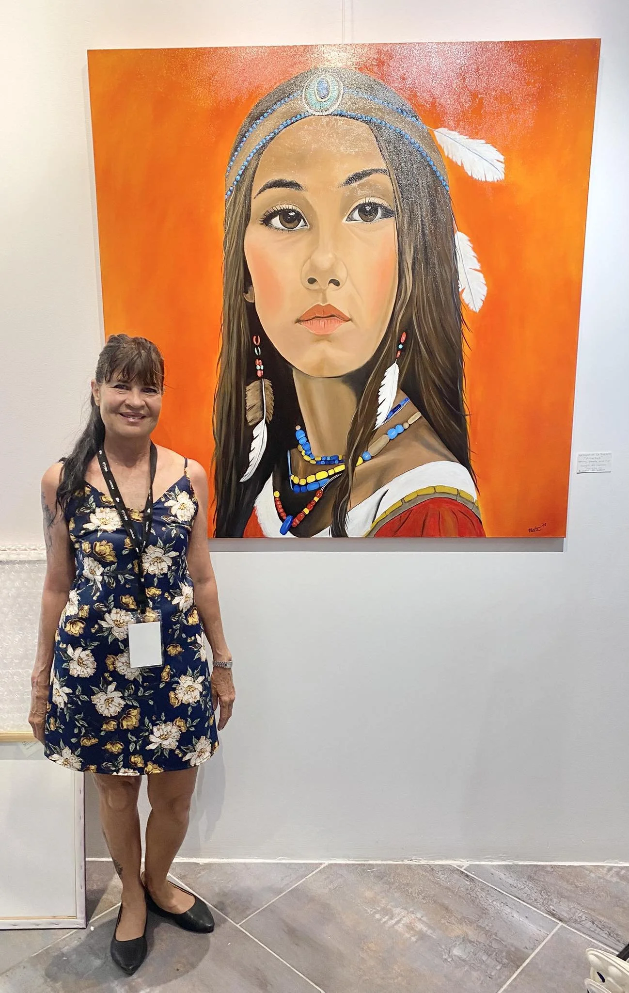 A woman standing in front of a large painting of a Native American woman with long dark hair, wearing traditional jewelry and feathers, with an orange background.