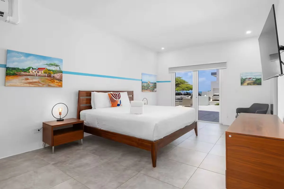A modern hotel room with a large bed, nightstand, and wall art, leading to a balcony with outdoor furniture and a parking lot view.