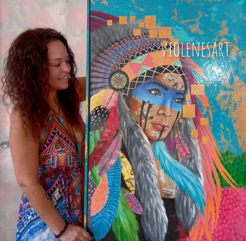 Woman with curly hair smiling next to a colorful mural of a Native American woman wearing a blue face paint and feathered headdress, with abstract shapes and vibrant colors in the background.