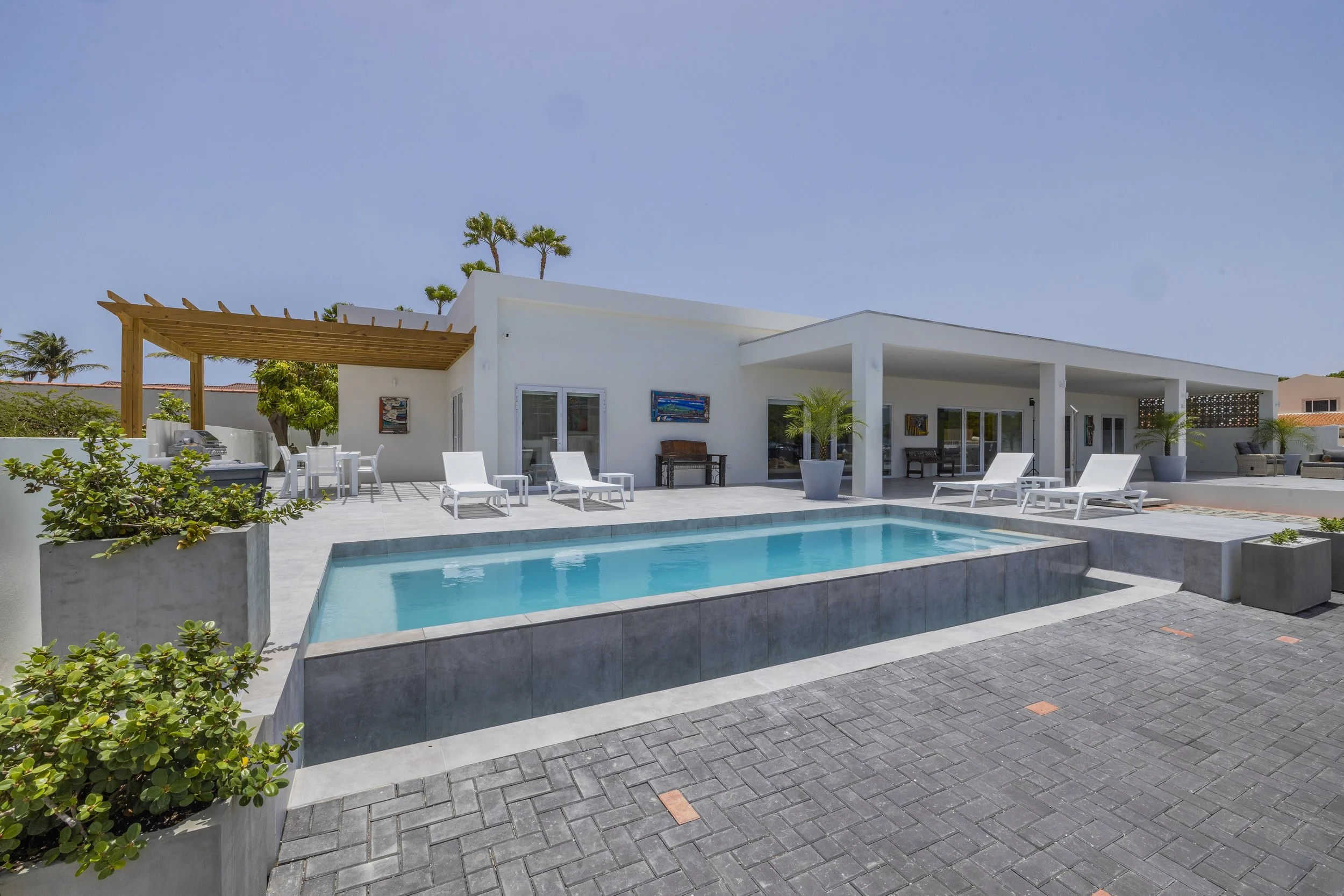 Modern white house with a backyard swimming pool, lounge chairs, potted plants, and outdoor seating under a clear blue sky.