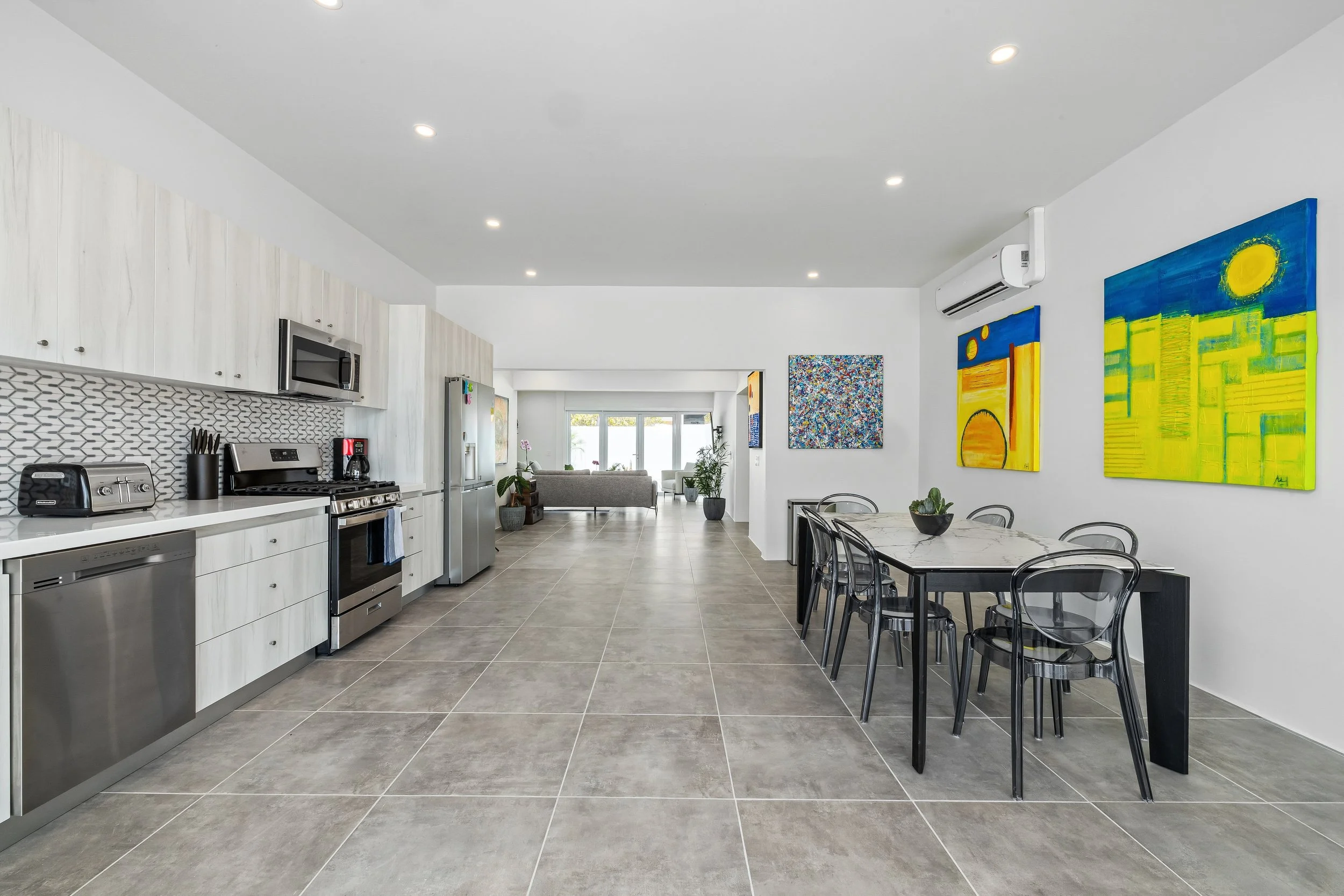 Open-plan kitchen and dining area with modern appliances, colorful wall art, and a view into a living room with a sofa and large windows.