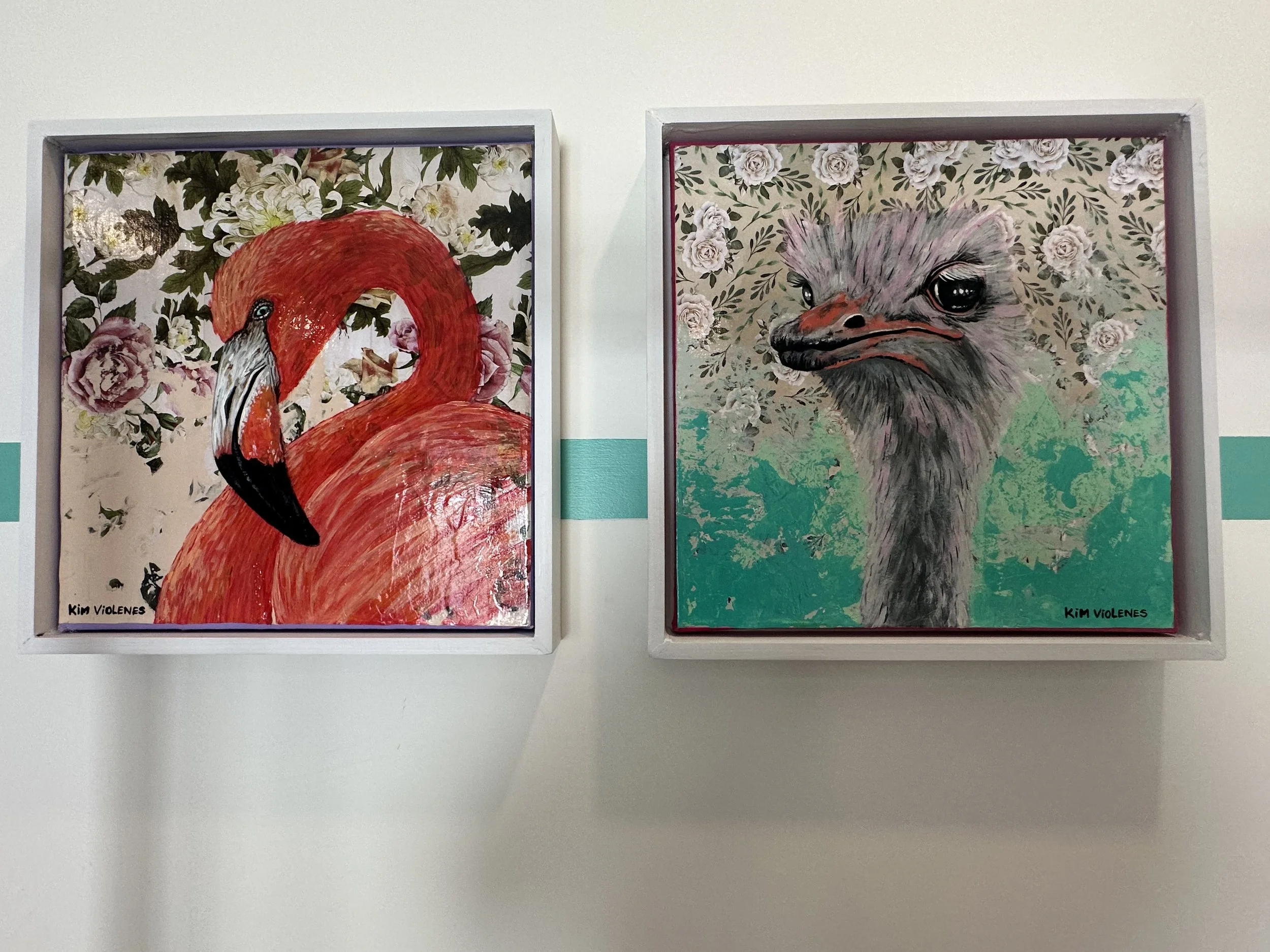 Two framed colorful animal paintings on a white wall. The left painting is of a pink flamingo with floral background. The right painting is of a bird with a floral background.