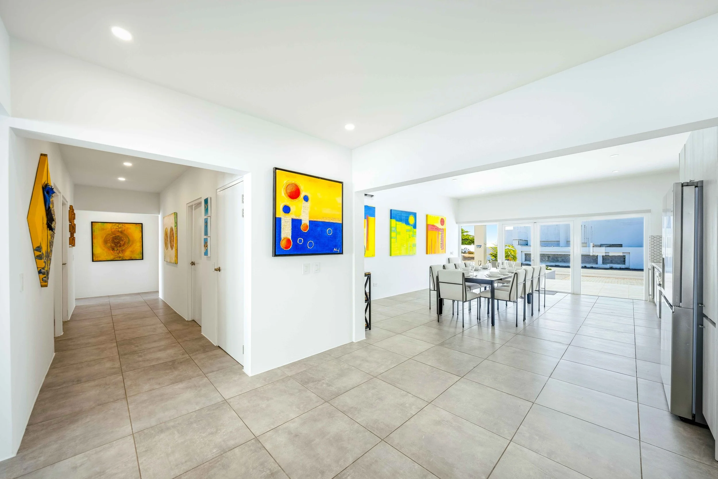 Bright, modern open-concept dining area with colorful abstract paintings on white walls, large windows and glass doors leading outside, and beige tiled floors.