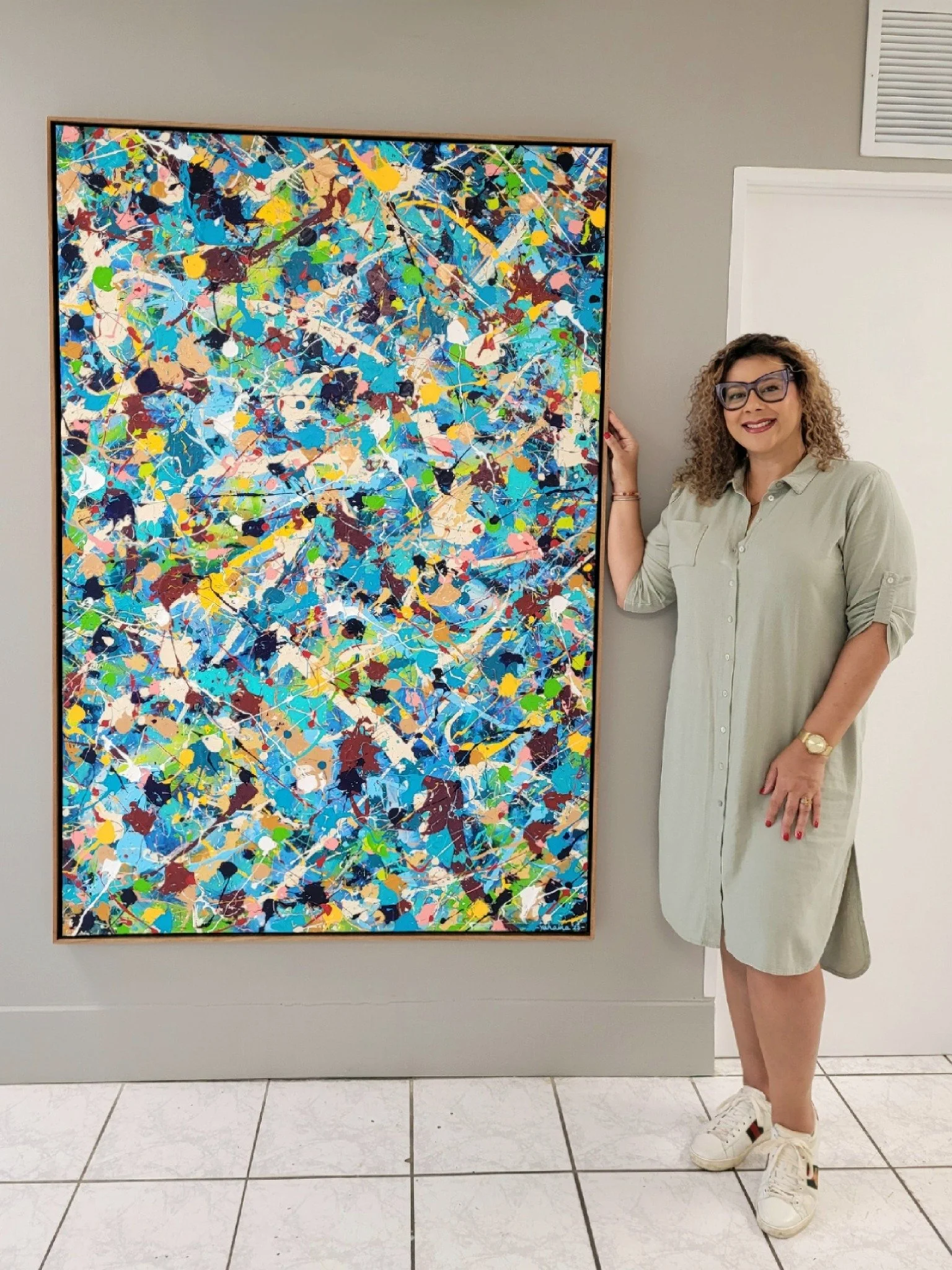 A woman with curly hair, glasses, and a beige dress standing next to an abstract colorful painting on a gray wall.